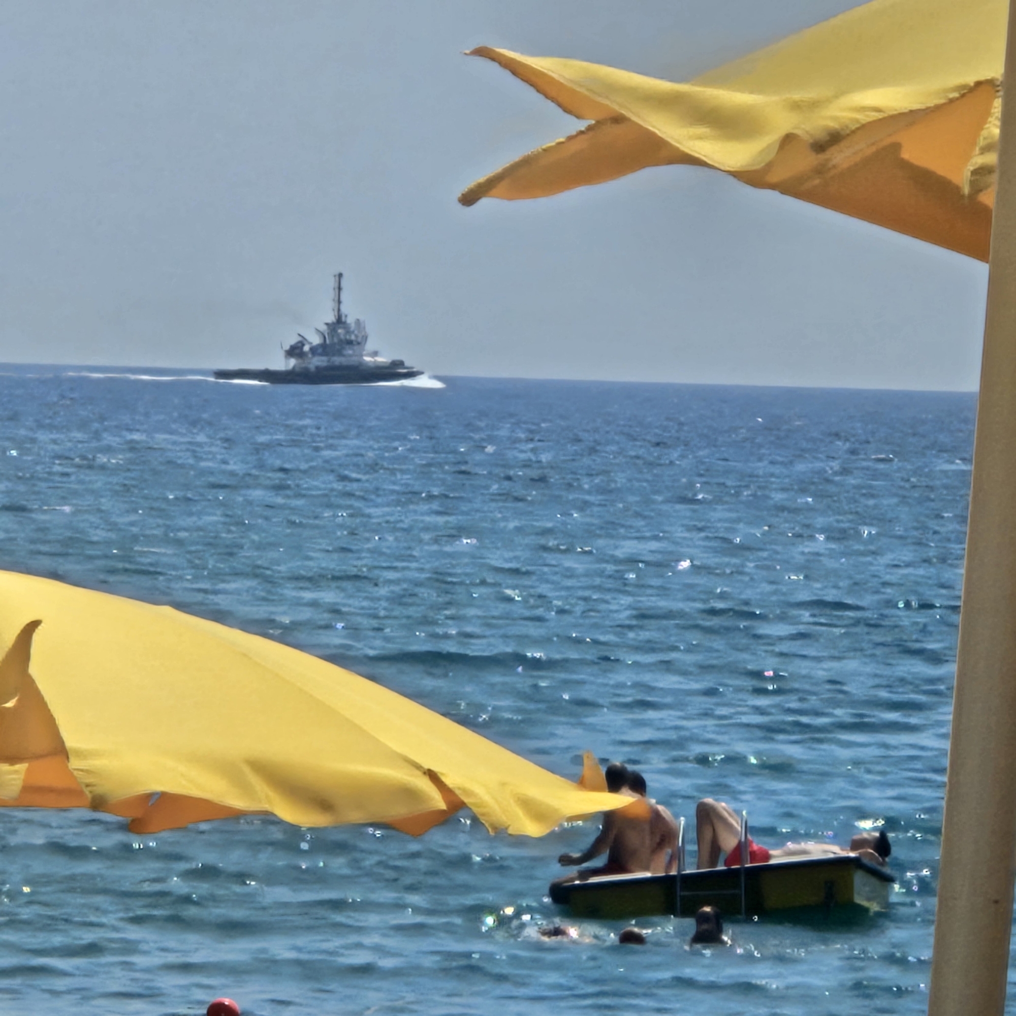 The image depicts a sunny day at the beach in Savona, Italy, with a tugboat in the distance on the blue sea. In the foreground, parts of two yellow umbrellas are visible, one on the left and one on the right, obscuring the sky and a portion of the water. In the water, several people are present. Two individuals are on a small yellow raft; one is seated and appears to be talking, while the other is lying down. Other swimmers are visible in the water around the raft. The sea is choppy with sunlight glinting off the waves.