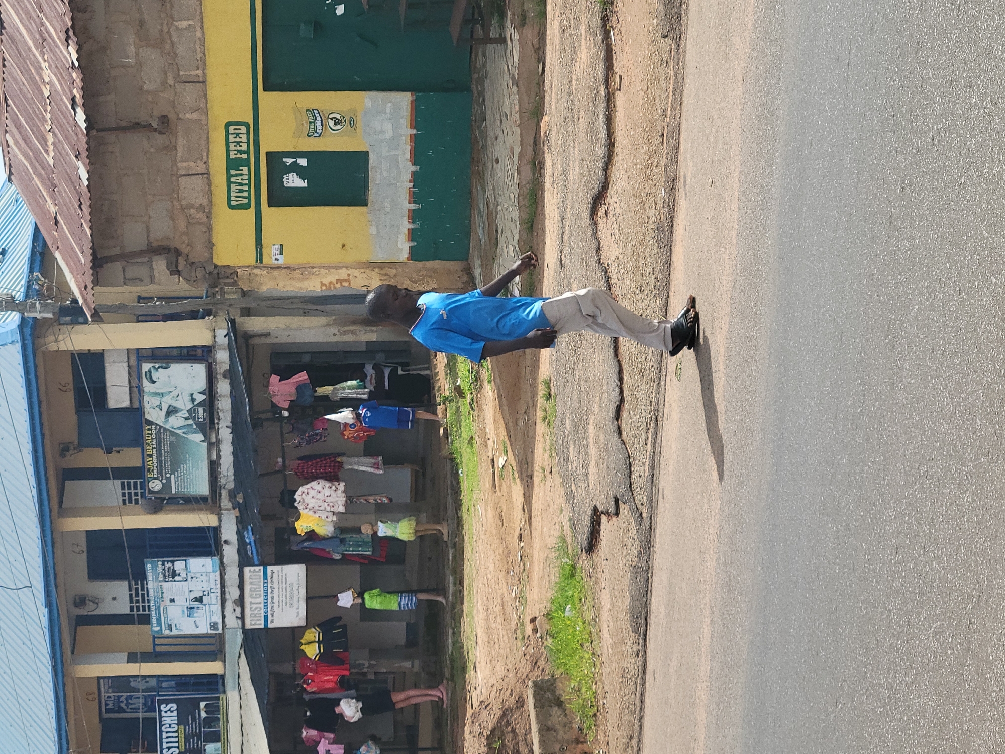 A boy walks on the side of a paved road in Jos, Nigeria. He is wearing a blue t-shirt and grey pants. In the background, there are shops with clothes hanging outside for sale. One shop has a sign that says "VITAL FEED". The buildings are constructed of concrete blocks and corrugated iron roofs. The road is in poor condition with patches of asphalt missing.