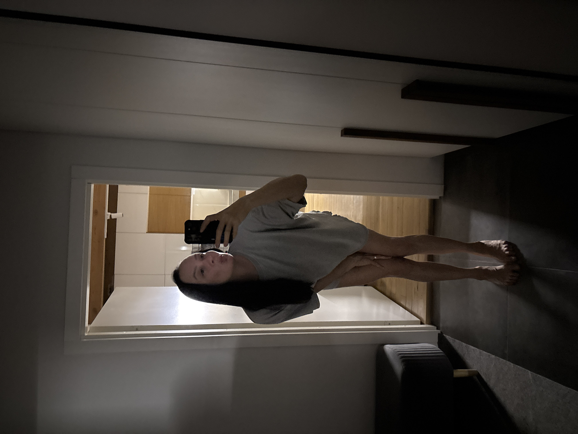 The image displays an indoor mirror selfie of a female subject in a residential environment. The woman has long, straight dark hair and is wearing a loose-fitting, light gray t-shirt. She is barefoot, with her left hand positioned near her upper thighs, obscuring them. Her right hand holds a dark-colored smartphone, used to capture the image, and her lips are pursed. A ring is visible on her left ring finger. The flooring in the immediate foreground consists of dark rectangular tiles. To the left, a dark gray upholstered bench is partially visible. Behind the subject, an open white doorway leads to a brighter room with light wooden flooring, white tiled walls, and light wooden cabinetry, indicative of a kitchen. The specific location within the city of Chaikovsky, Russia, is not discernible from the visual evidence presented.