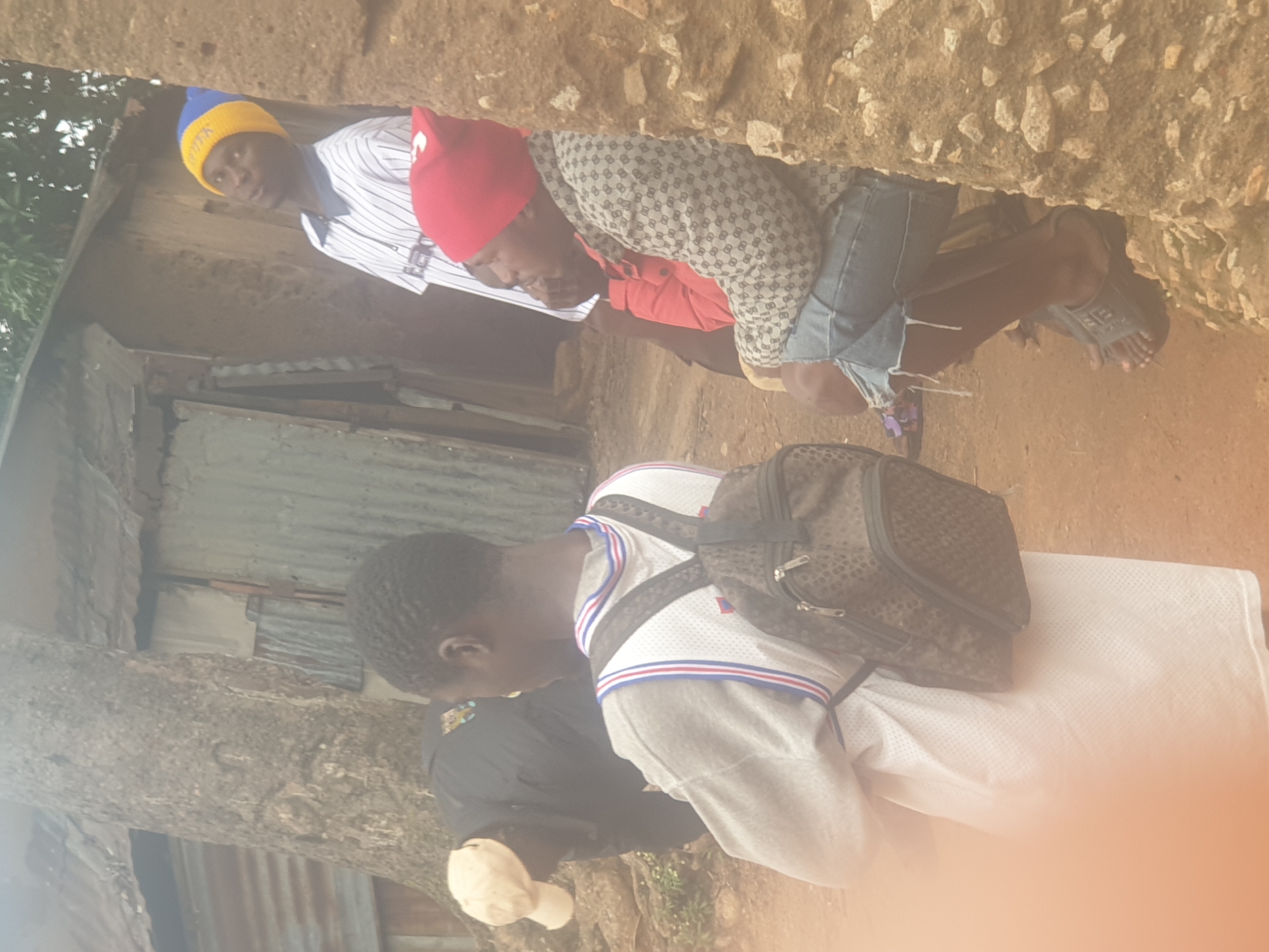 The image depicts a group of individuals outdoors, with a focus on a young man in the foreground wearing a white t-shirt with blue and red trim and a patterned backpack. To his right, a person in a patterned jacket and denim shorts, wearing a red beanie, is seated. Behind them, a man in a striped shirt and a blue and yellow beanie looks upwards. Another individual, partially obscured, is wearing a light-colored cap and a dark jacket, with their back to the camera. The environment appears to be rustic, with a large tree trunk and a corrugated metal structure in the background. The ground is reddish-brown dirt. The scene does not provide any specific indicators to determine its location within Washington, United States of America.