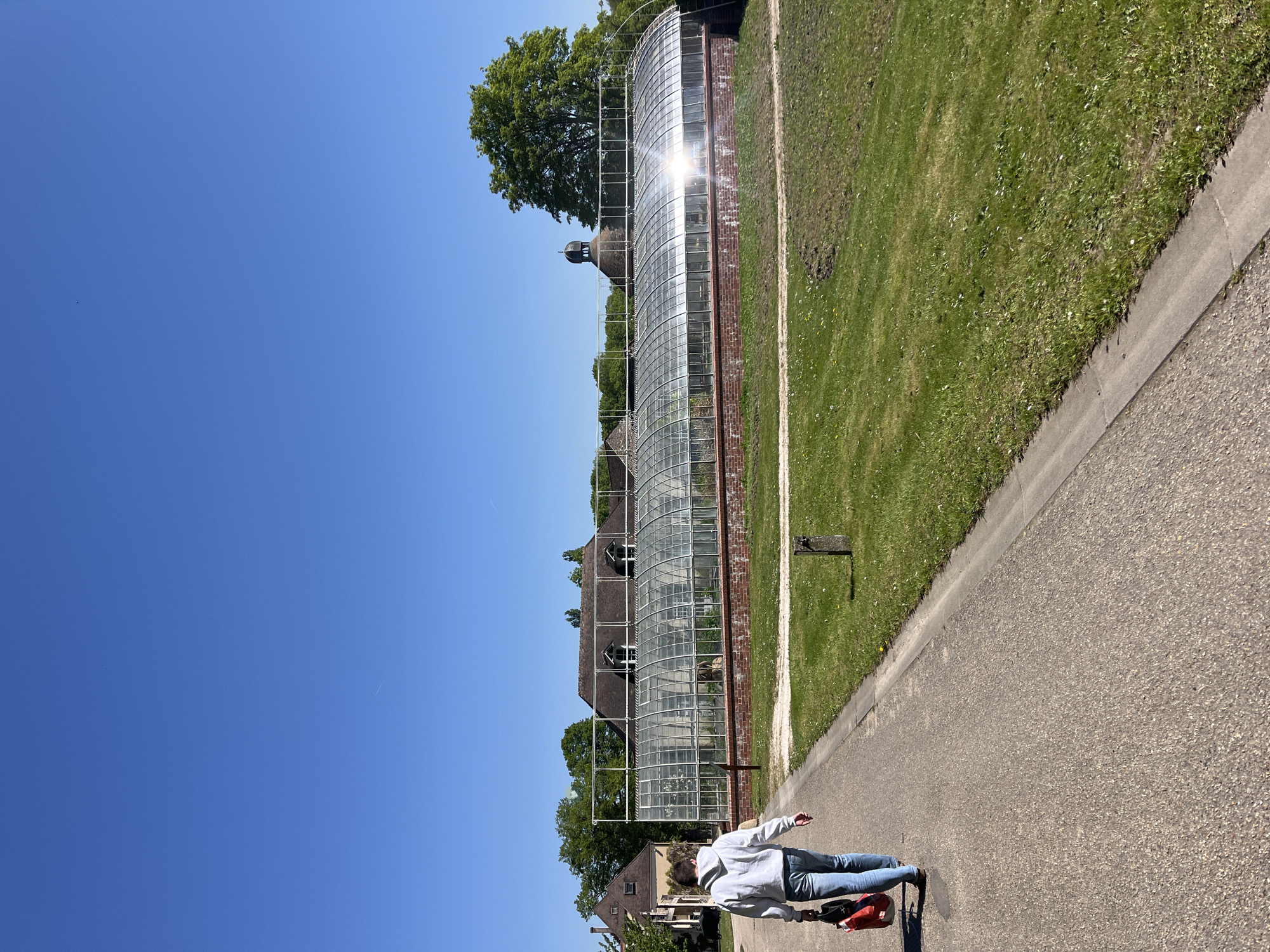 The image shows a person walking on a paved path in Saint-Rémy-lès-Chevreuse, France. The person is wearing a light-colored hoodie and blue jeans, and they are pulling a red bag on wheels. To the right of the path is a grassy area. In the background, there is a large glass greenhouse-like structure with a brick base. Adjacent to the greenhouse is a building with a dark roof. The sky is clear and blue. Trees are also visible in the background.