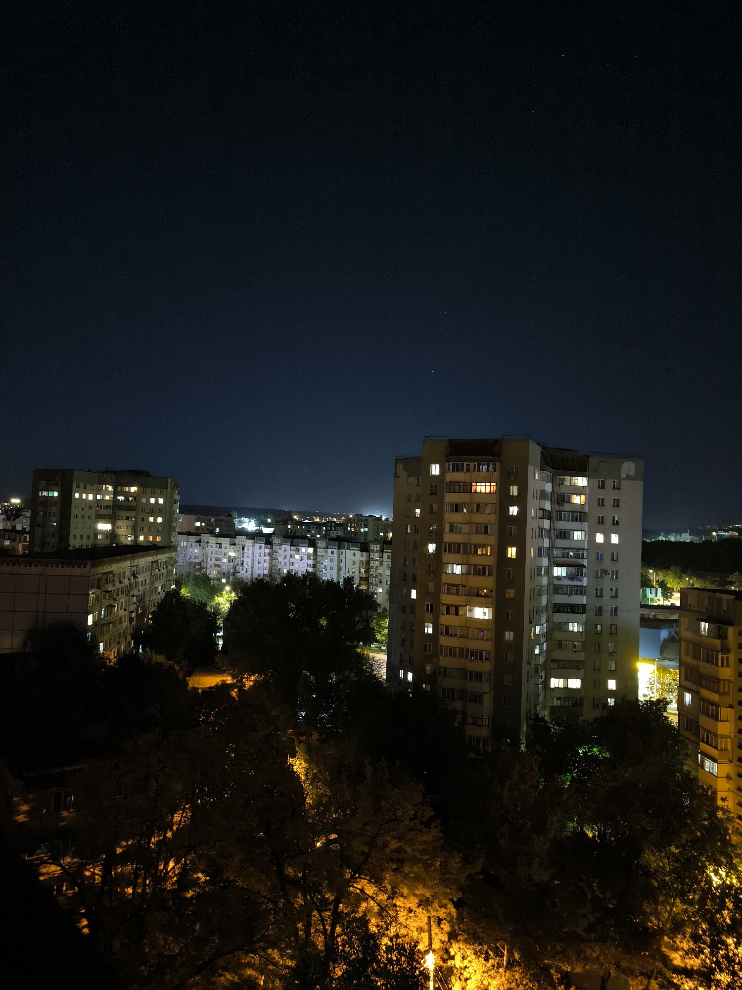 This is a night-time, high-angle, exterior, full shot of a residential area in Chișinău, Moldova. The foreground is dominated by the dark silhouettes of trees, with some branches illuminated by warm, artificial light sources below. Beyond the trees, several multi-story apartment buildings are visible. Many windows are lit, indicating occupied apartments. The buildings are of varying heights and architectural styles, some appearing older and more weathered, while others look more modern. The sky above is dark and clear, with a few faint stars visible. In the distance, the lights of the city extend towards the horizon, creating a glowing urban landscape. The overall impression is one of a typical urban night scene in a residential district.