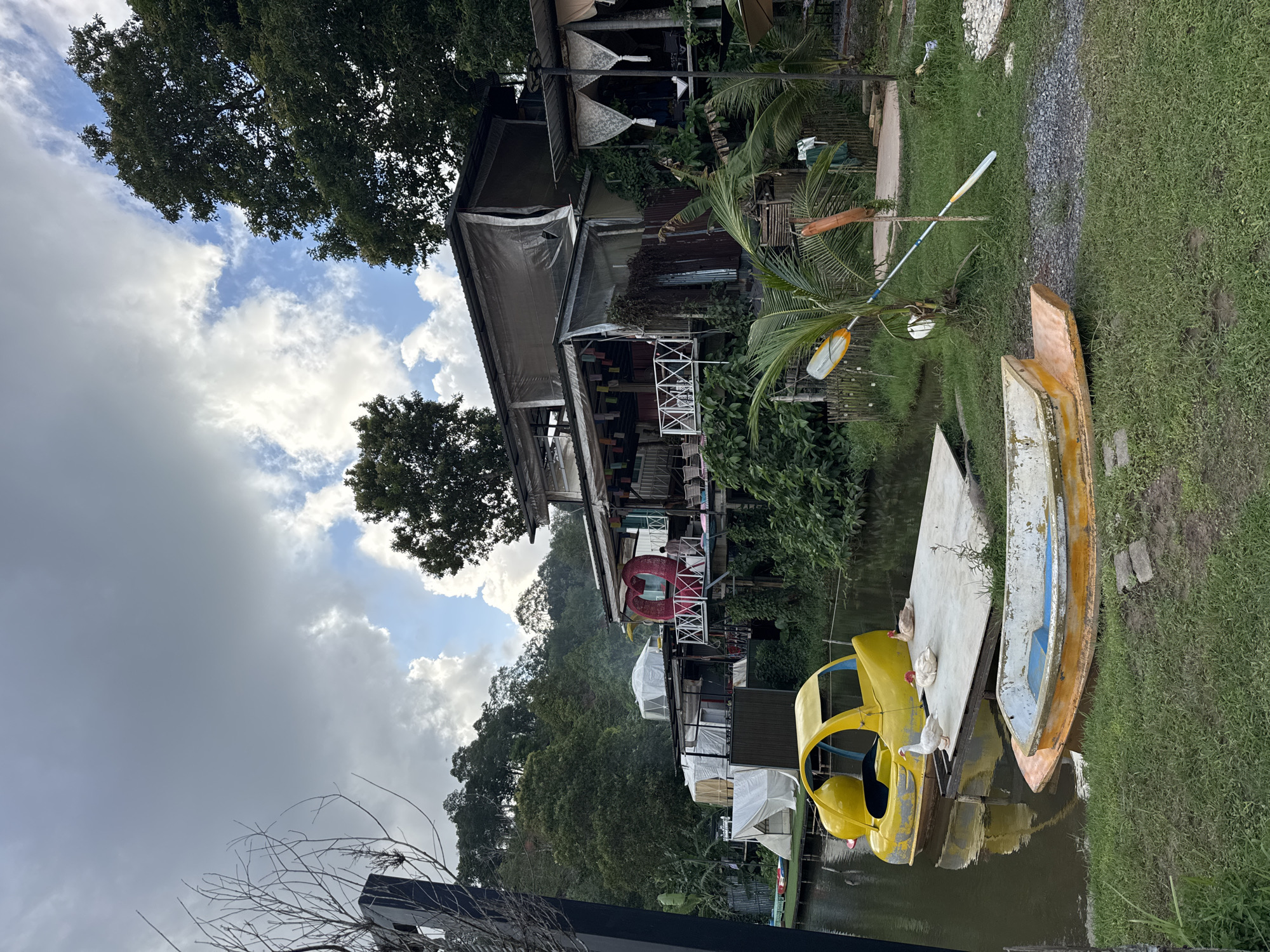 The image depicts a waterside establishment with multiple levels, situated in Kathu, Thailand. The architecture is eclectic, featuring a main structure with open-air seating areas covered by awnings and tarps. A prominent red heart sculpture is visible on a balcony. In the foreground, a yellow novelty boat, resembling a car, floats on the water, alongside a white platform on which three ducks are resting. Another boat, yellow and white with blue interior, is beached on the grassy bank. The scene is surrounded by lush greenery and mature trees, with a cloudy sky overhead. The overall impression is that of a tranquil, recreational lakeside resort or cafe.