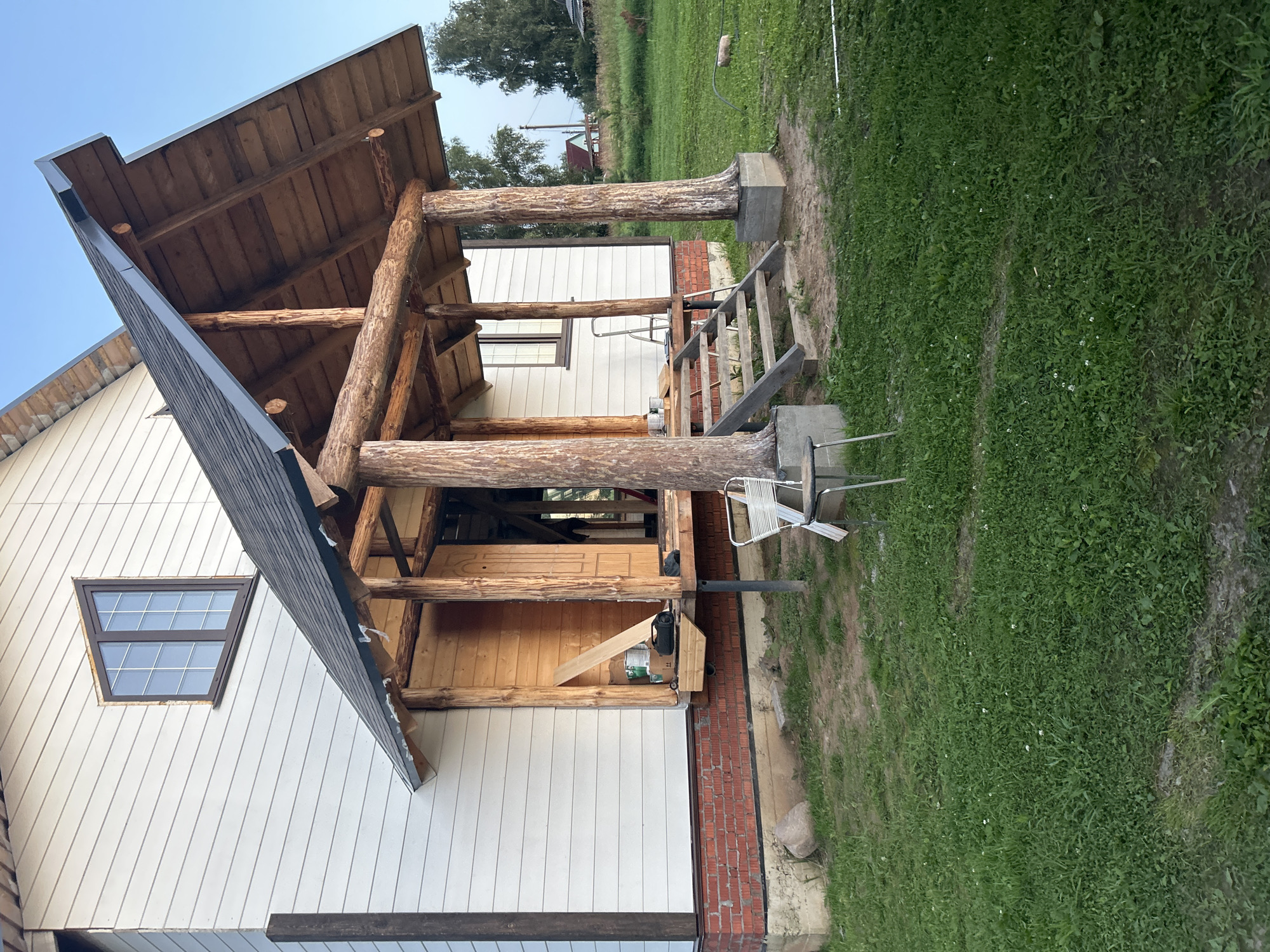 This image depicts the exterior of a house under construction in Vereiki, Russia. The house is clad in white horizontal siding with a visible red brick foundation. A wooden porch with a dark shingled roof is a prominent feature. The porch is supported by thick, natural logs, giving it a rustic appearance. Wooden stairs lead up to the porch, and a ladder is visible leaning against the house. Various construction materials and tools, including cans of paint and wooden planks, are scattered on and around the porch. The surrounding environment is a grassy yard with some trees in the background. The sky is clear blue, suggesting fair weather. No people are visible in the image.