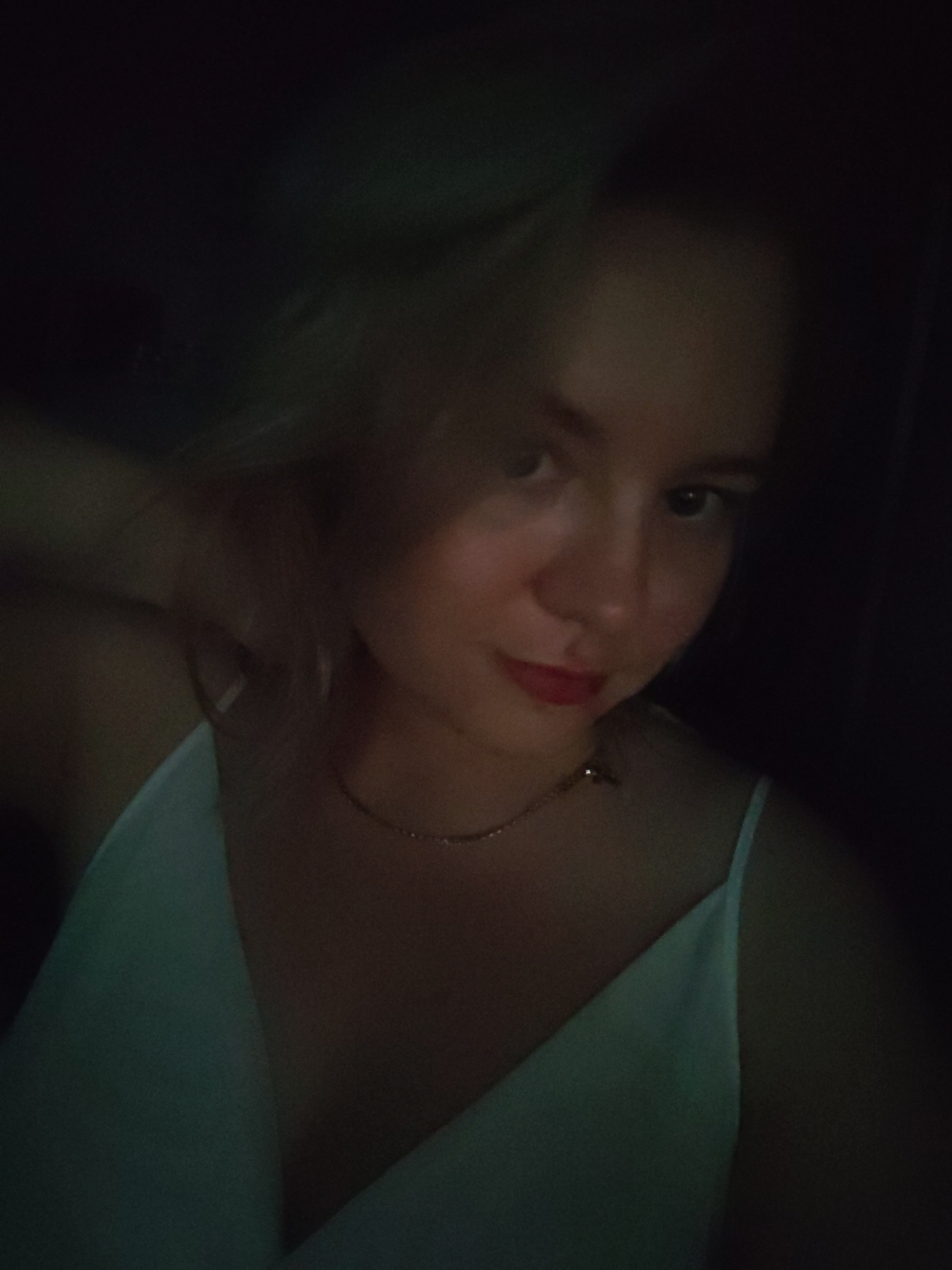 A young woman is pictured in a close-up, self-taken photograph. She has fair skin, blonde hair, and is wearing a light-colored, possibly white, spaghetti strap dress. A delicate gold necklace is visible around her neck. Her lips are slightly parted and appear to be colored with lipstick. The background is dark and out of focus, suggesting an indoor setting with low lighting. No specific location in Izhevsk, Russia can be identified from the image.