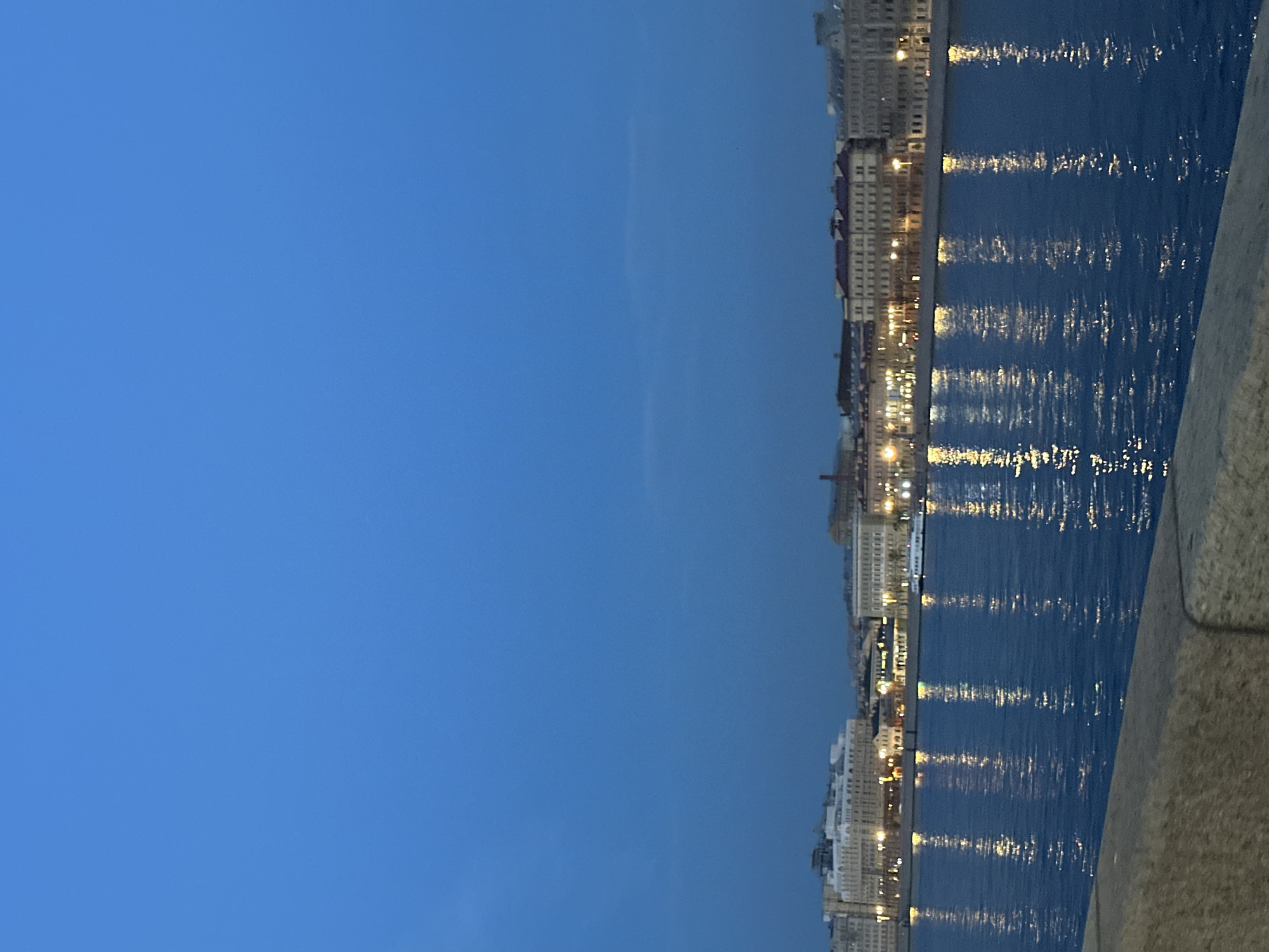 The image depicts a waterfront cityscape at dusk. A wide body of water, likely a river, occupies the lower half of the frame, reflecting the lights of buildings on the opposite bank and the soft glow of the twilight sky. The sky is a deep, clear blue, transitioning into a lighter hue near the horizon.
On the far side of the water, a line of historic buildings stretches across the view. These structures appear to be made of stone or brick, with multiple stories and architectural details common to European cities. Many windows are illuminated, creating warm, yellow reflections that shimmer on the water's surface. A white boat is visible on the water, positioned in front of the buildings.
In the foreground, the edge of a stone embankment or walkway is visible, providing a vantage point for the photographer. The overall mood is tranquil and serene, characteristic of a city at the end of the day. The scene is devoid of any prominent human figures or active events, focusing instead on the architecture and the interplay of light and water.