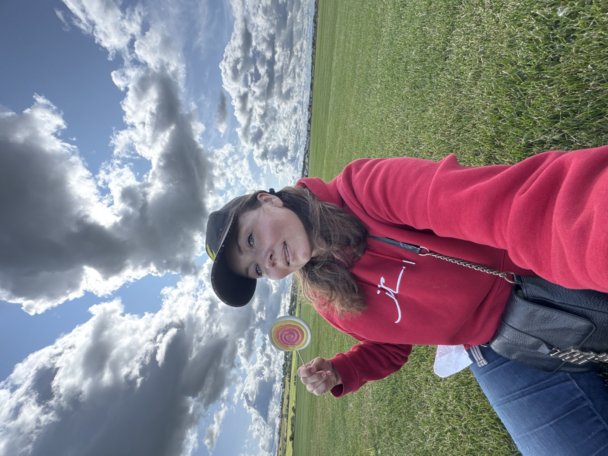 Innolovo, Russia - A woman wearing a red hoodie, blue jeans, and a baseball cap sits in a grassy field under a cloudy sky. She is holding a large, colorful lollipop in her right hand. A black purse hangs from her left shoulder. In the background, there is a distant treeline and a few houses.
