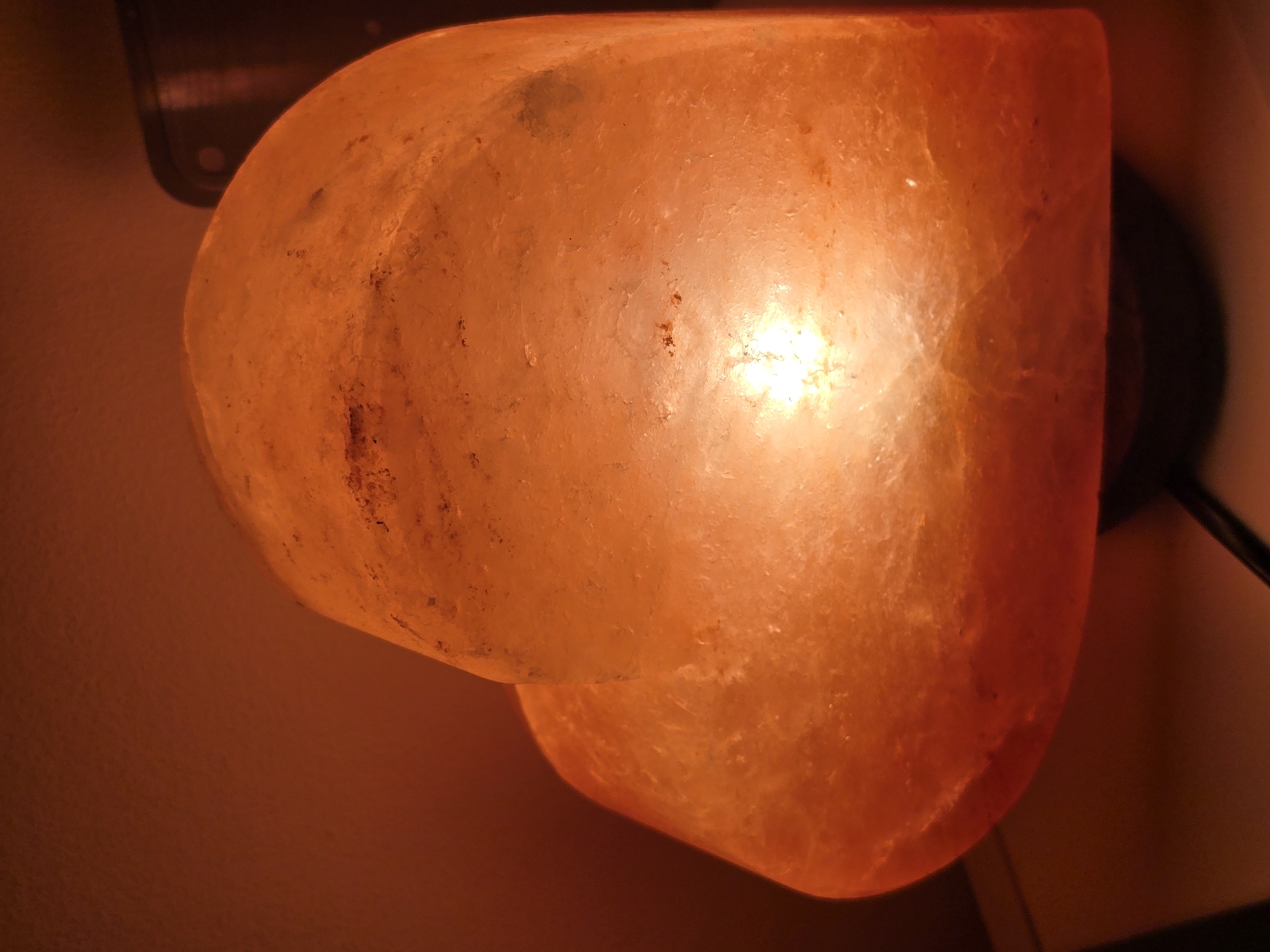The image shows a close-up of a heart-shaped Himalayan salt lamp, illuminated from within, casting a warm, orange glow. The surface of the lamp is rough and crystalline, with visible natural imperfections and darker mineral inclusions. The background is out of focus, a warm, reddish-orange hue, suggesting a dimly lit interior. The base of the lamp and a portion of a black electrical cord are visible at the bottom. The scene appears to be a personal item in a domestic setting. There is no indication of the city of Golyanovtsi, Bulgaria.