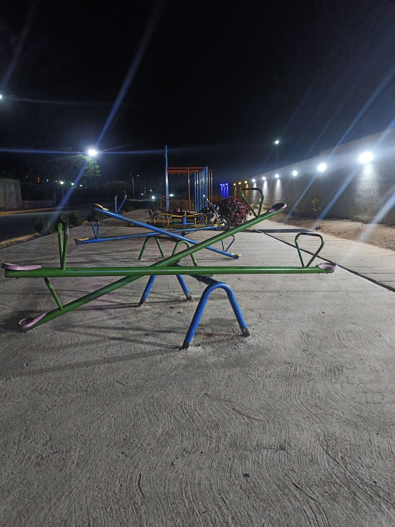 The image depicts a deserted outdoor playground area at night in Tinaquillo, Venezuela. In the foreground, two metal seesaws are prominently positioned on a patterned concrete surface. One seesaw is green with pink seats and handles, resting with one end low to the ground. Directly behind it, another seesaw is blue with yellow seats and handles, tilted upwards.
In the mid-ground, additional playground equipment, including a blue and orange metal climbing or swing structure, is partially visible. A dark motorcycle is discernible behind the blue seesaw. To the left, a dark asphalt road is visible, bordered by low vegetation and illuminated by distant streetlights. To the right, a light-colored, textured wall is brightly illuminated by a series of at least five white floodlights mounted along its top edge, casting distinct light streaks across the frame. Some reddish-purple shrubbery is present near the illuminated wall and the playground equipment. The sky is dark, indicating nighttime, with multiple streetlights and artificial lights providing illumination, resulting in prominent lens flares or light streaks from several sources. No individuals are present in the scene, and no activity is occurring.