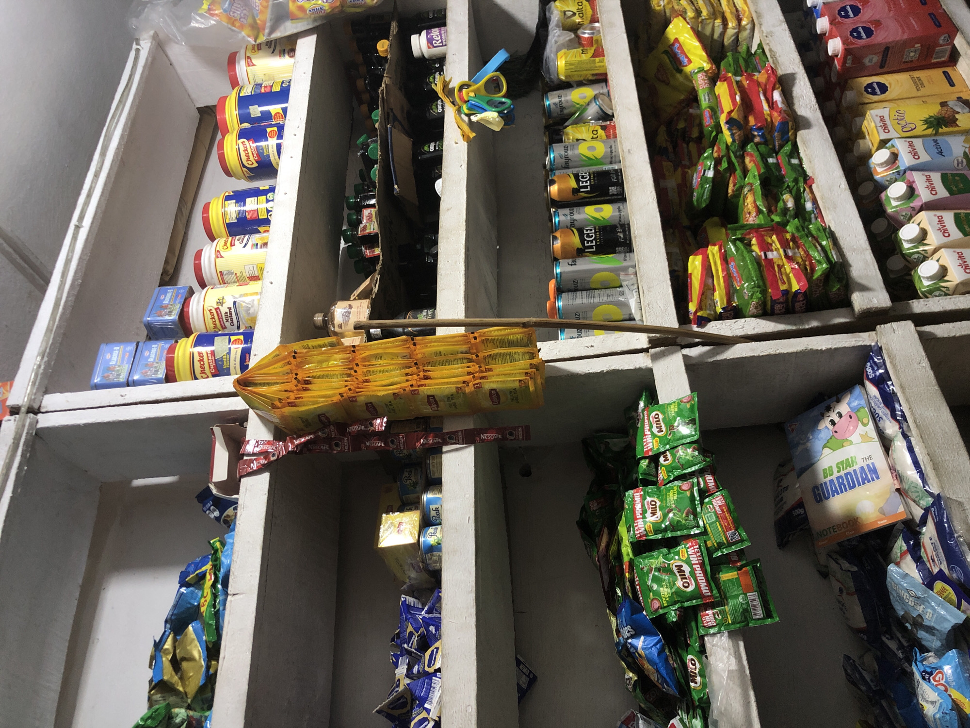The image depicts a cluttered interior scene of a small retail shop, likely a convenience store or a local market stall, in Ado, Nigeria. The shelves are densely packed with various packaged goods.
On the top shelf, several blue boxes labeled "Glucose-N" are stacked, along with yellow and white tubs of what appear to be food products, some with red lids and labels that include "Checkers Custard". To the right, a partially visible plastic bag contains more yellow-packaged items.
The middle shelf holds a variety of items including packets of what seem to be dried foods or snacks, some in blue and gold packaging. A yellow bag filled with small, individually wrapped items hangs from the shelf. Further to the right, a row of beverage cans is visible, some with labels indicating "Fayrouz" and "Legen". There are also bottles of what appear to be condiments or sauces, some in dark green bottles with black caps, and a few smaller bottles with yellow caps on the far right.
The lower shelf is also filled with various products. Prominently displayed are numerous green packets labeled "Milo", suggesting a focus on breakfast or snack items. Below these, there are more packaged goods in blue and white wrappers, and a book or notebook with a cartoon cow and the label "Guardian". Further to the right, there are rows of cartons containing beverages, including "Chivita" branded drinks in different flavors, and a red carton labeled "Malta".
The overall environment suggests a typical small business operation, with goods arranged for easy access and display. The lighting is somewhat dim, contributing to the cluttered appearance. The scene is static, with no people or active interactions visible.