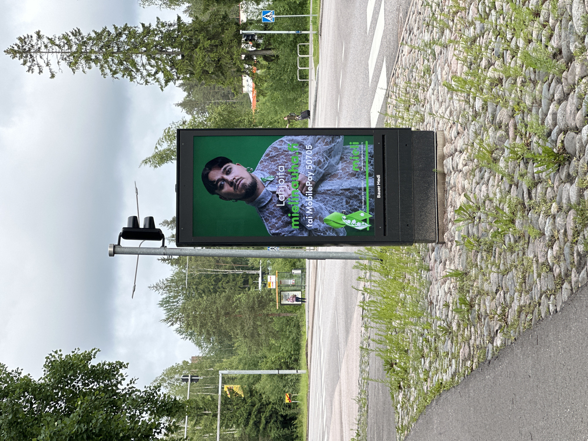 This is a medium shot of a digital billboard on a cloudy day. The billboard displays an advertisement featuring a person and text. The person in the advertisement is looking directly at the camera and is wearing a sheer, white lace shirt. Text on the billboard includes "Lahjoita mistinauha.fi", "tai MobilePay 50705", and "Bauer Media". The billboard is situated on a sidewalk paved with cobblestones, with green grass growing between the stones. To the left of the billboard is a paved road with a crosswalk. In the background, there are trees, a traffic light, and a bus stop with a single person waiting. Further in the distance, there are buildings and more trees. The sky is overcast.