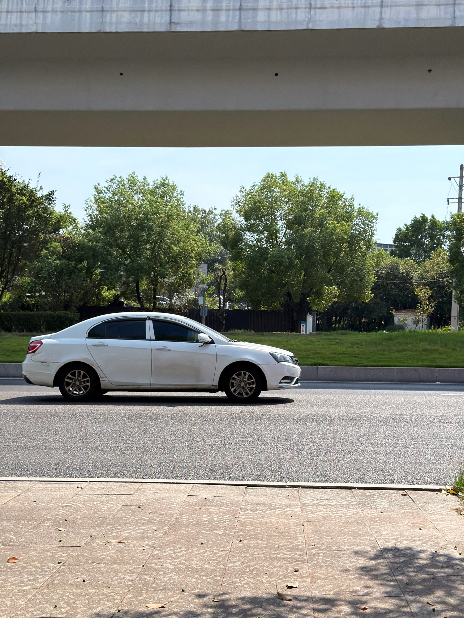 A white sedan is driving on a paved road in Jiangjin, China. The car is in the middle lane of a multi-lane road, with a sidewalk to the left and a grassy area with trees to the right. An elevated structure, possibly an overpass or bridge, is visible in the upper portion of the image, casting a shadow on the road. The sky is clear and blue. The car appears to be in motion, with its wheels turned slightly. There are no pedestrians or other vehicles immediately visible. The scene suggests an urban or suburban environment.