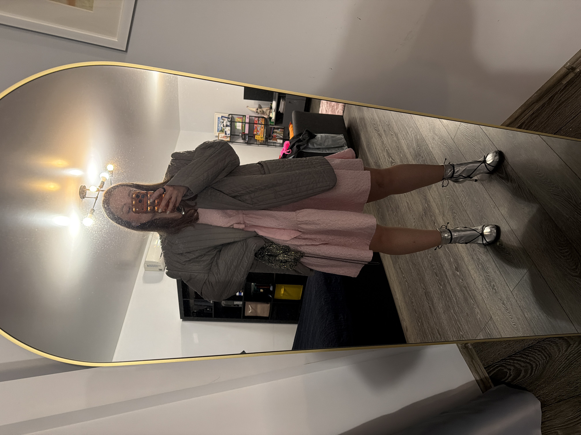A person is taking a photograph of themselves in a mirror. The person is wearing a pink dress, a gray jacket, and silver shoes. The mirror has a gold frame. The background includes a shelf filled with items, a black couch, and a wooden floor. A painting is hanging on the wall, and a chandelier is reflected in the mirror. The location in the city of Moscow, Russia is not discernable.