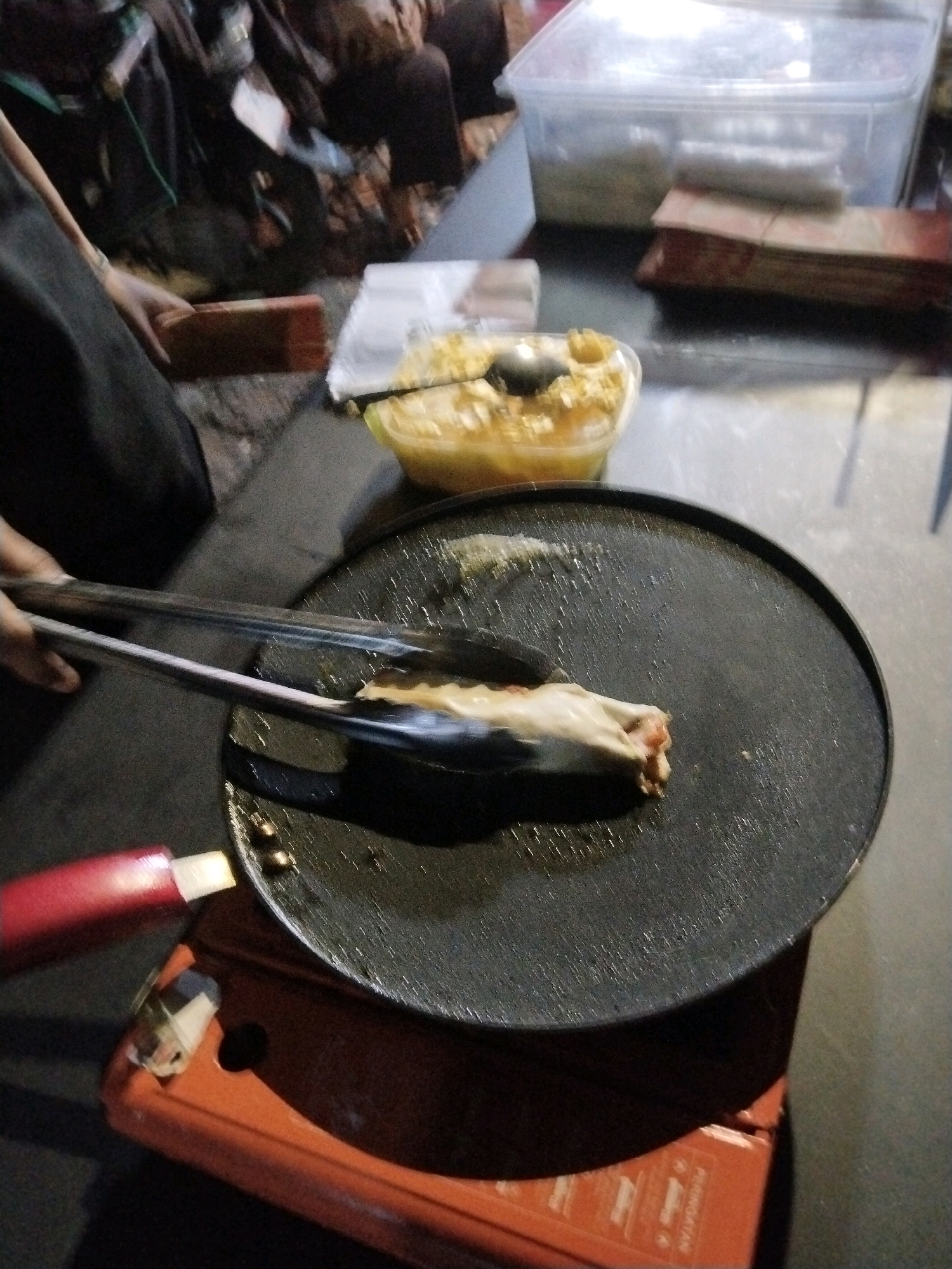 The image shows a close-up of food preparation, likely in an outdoor or semi-outdoor setting given the ambient lighting. A person's hand, wearing a dark sleeve, is using metal tongs to handle a piece of food on a black griddle. The food appears to be wrapped in a thin, light-colored dough and is being flipped or cooked. The griddle is placed on an orange portable stove. In the background, there is a clear plastic container filled with a yellow, chunky substance, possibly a sauce or topping, and other containers and stacked items that are out of focus. The scene suggests street food preparation or a casual cooking event, possibly in Slawi, Indonesia, given the provided context.