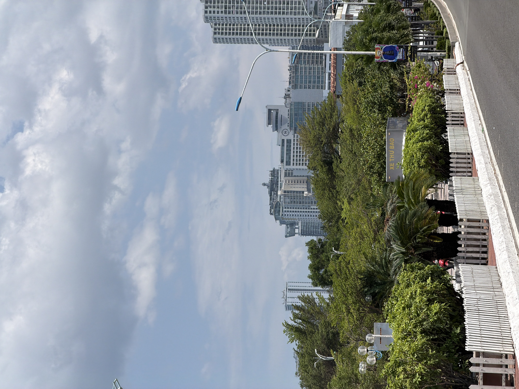 This media file captures an urban street scene in Bac Nha Trang Ward, Vietnam, under a partly cloudy sky during daylight hours. The image is oriented vertically with the sky on the left and the ground on the right.
In the foreground, a paved road with a white curb runs along the right side of the frame. Adjacent to the road is a white picket fence, behind which lies a lush, green median or park area filled with various trees, bushes, and tropical plants, including palm trees. Modern streetlights with blue-tipped fixtures are interspersed within this greenery. A dark sign with white text, "XE THU BAO," is clearly visible within the vegetation. Further down, another banner or sign features an illustration and partially visible English text, "NEW RUNNING CHALLENGE."
The midground and background are dominated by numerous contemporary high-rise buildings with many windows, indicating a developed urban landscape. One building has a distinct circular clock-like feature on its facade, and another shows text that appears to be "HOAN CAO." The sky above is largely covered with light grey and white clouds. No people or specific activities are discernible in the image, depicting a static city environment.
