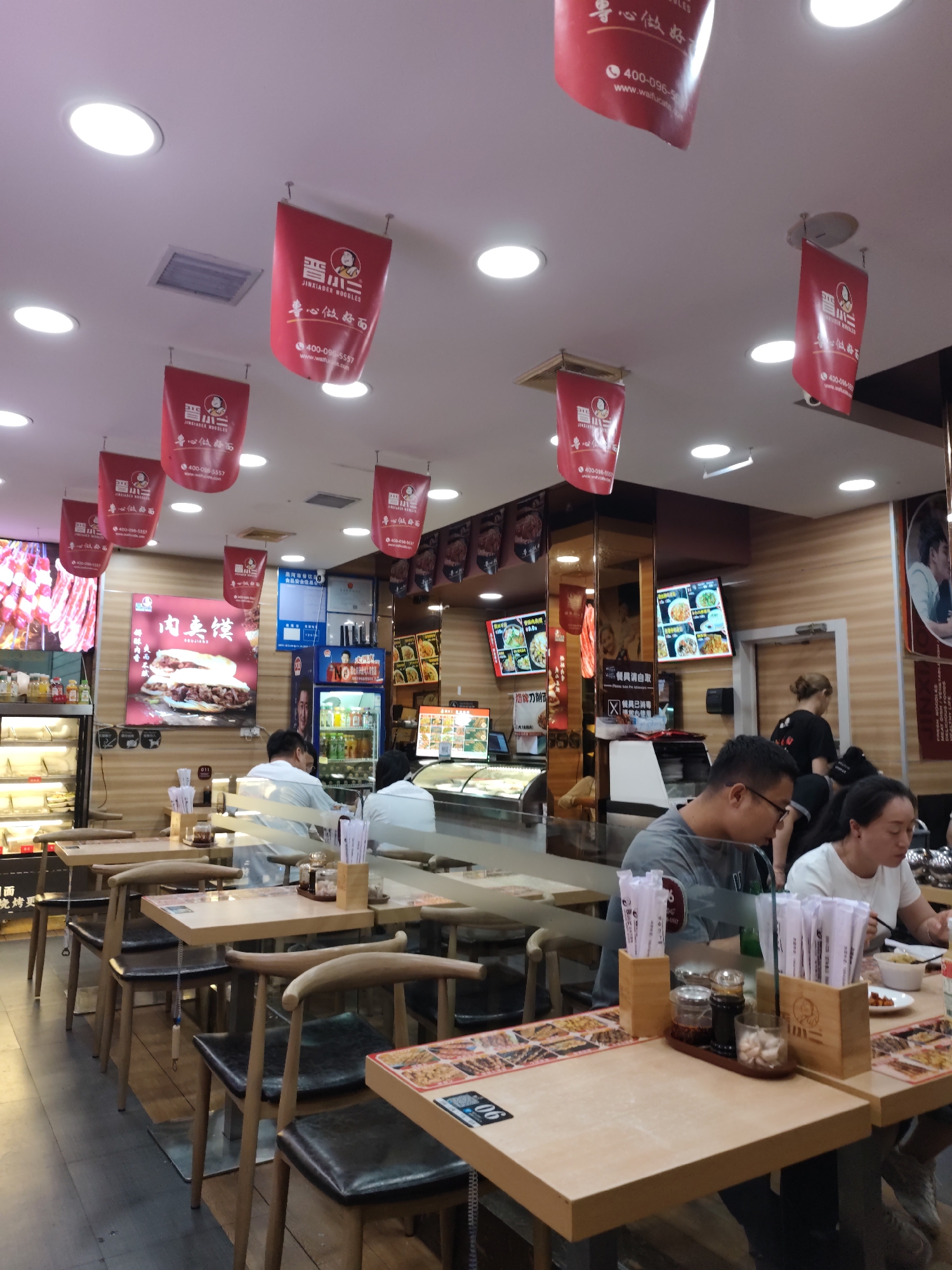 The image shows the interior of a restaurant in Aihui, China. The restaurant appears to be a casual dining establishment, possibly a noodle shop, given the signage and the hanging banners. The decor is simple with wooden tables and chairs. Several red banners with Chinese characters and logos are hanging from the ceiling. In the foreground, two individuals are seated at a table, one man wearing glasses and a grey t-shirt, and a woman in a white t-shirt. They are in the process of eating. The table is set with chopsticks, napkins, and condiments. In the background, other patrons and staff are visible. The restaurant has display cases with food items, a refrigerator stocked with drinks, and digital menu boards on the wall. The lighting is artificial, with circular ceiling lights illuminating the space. The overall atmosphere suggests a busy lunchtime or dinner service.