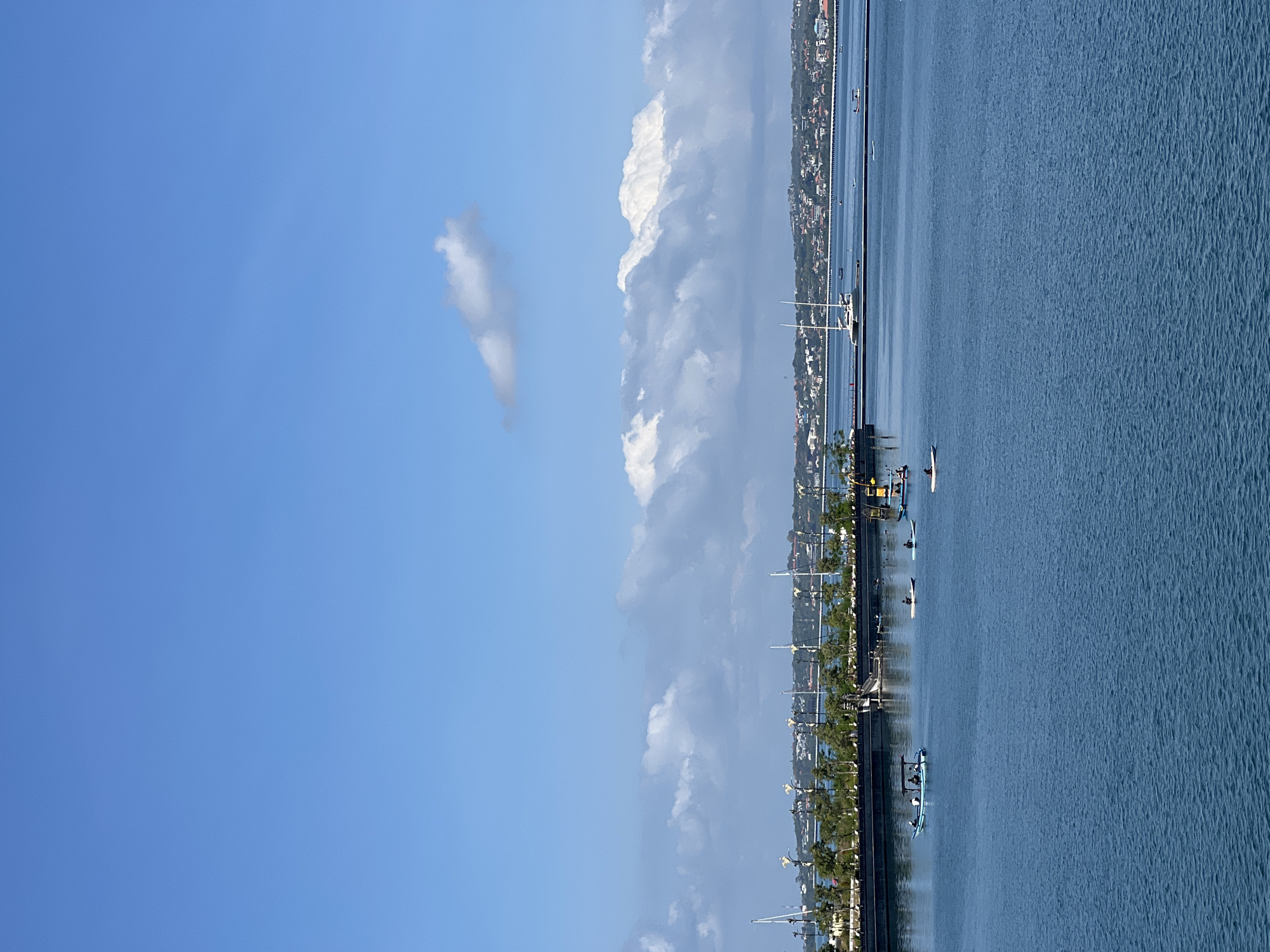 The image shows a view of the water in Kuta, Indonesia, with the coastline visible in the background. Several boats and paddleboarders are in the water. The sky is blue with scattered clouds. The weather appears clear. A row of trees lines the coast.