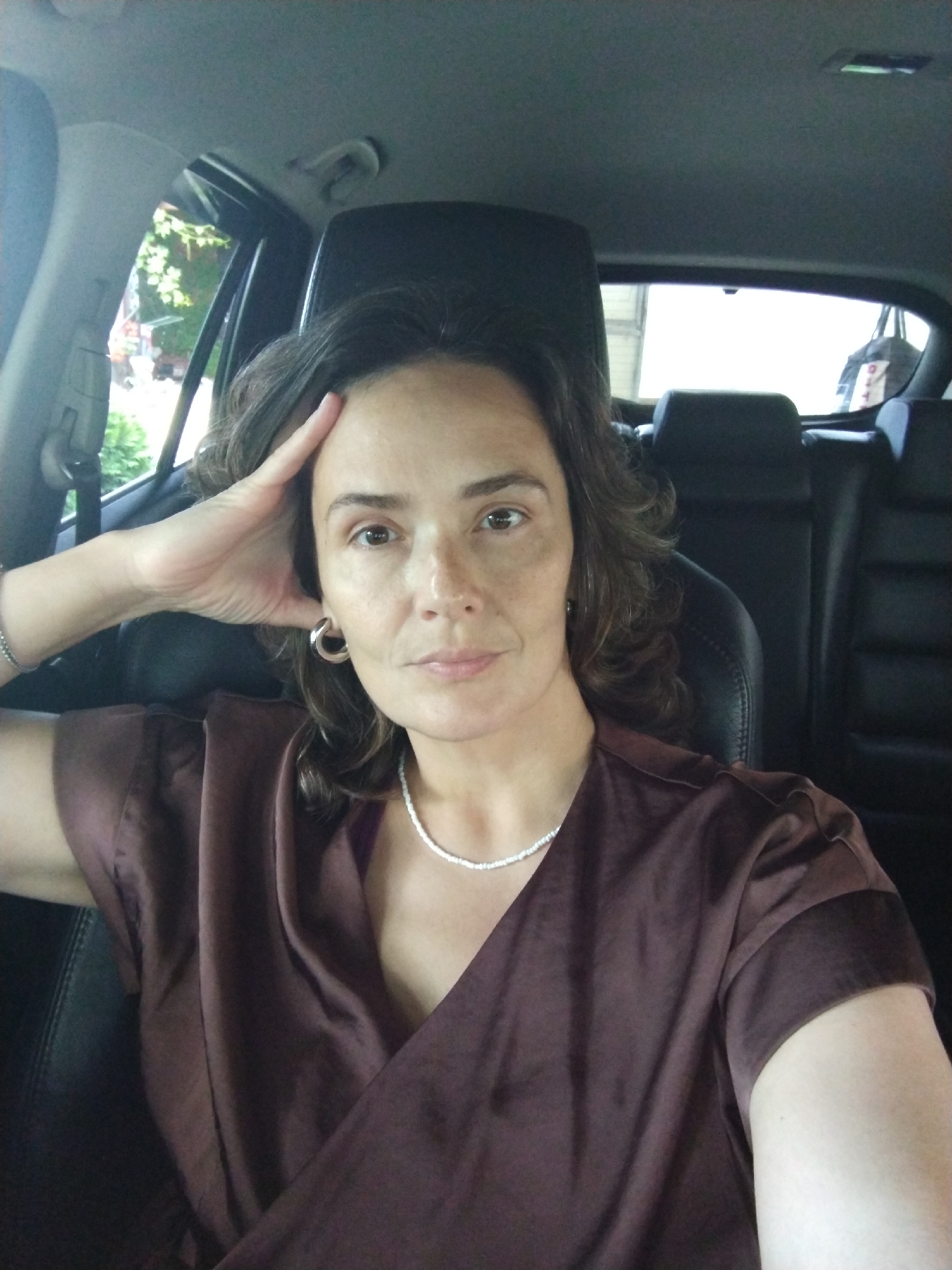 A woman is sitting in the driver's seat of a car, with her left arm resting on her forehead. She is wearing a dark brown satin top and a silver beaded necklace. The interior of the car, including black leather seats and headrests, is visible behind her. Through the car windows, greenery and trees are visible, suggesting an outdoor location. The scene appears to be a portrait taken inside a vehicle. The exact location within the city of Vinogradny, Russia cannot be determined from the image alone.