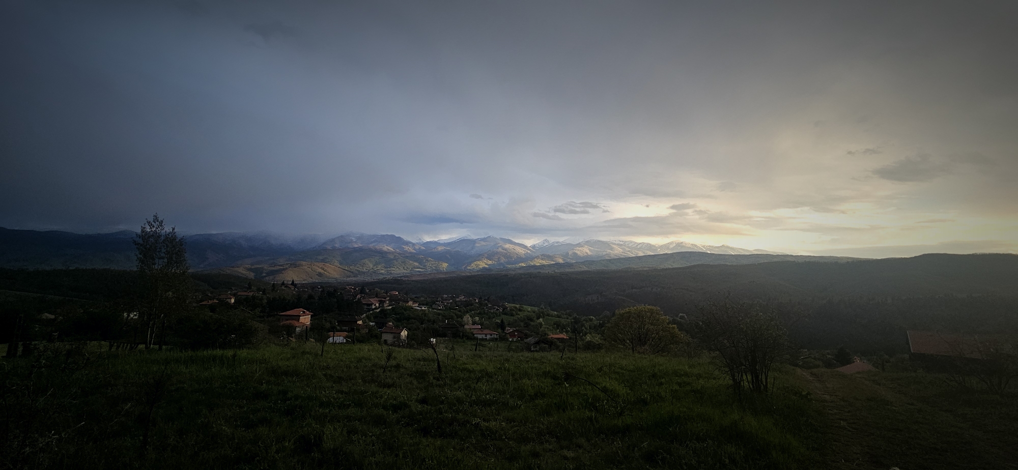 The image depicts a landscape in Gutsal, Bulgaria. In the foreground, a grassy field slopes downward, partially obscuring several houses with red roofs in the middle distance, suggesting a village. Beyond the village, a series of mountain ranges stretch across the horizon, with some peaks capped with snow. The sky is overcast, with a dark, stormy area looming over the left side of the frame and a lighter, brighter area to the right, possibly indicating either dawn or dusk. There is a small tree on the left and a small shrub on the right side. The lighting in the field is dim due to the cloud cover.