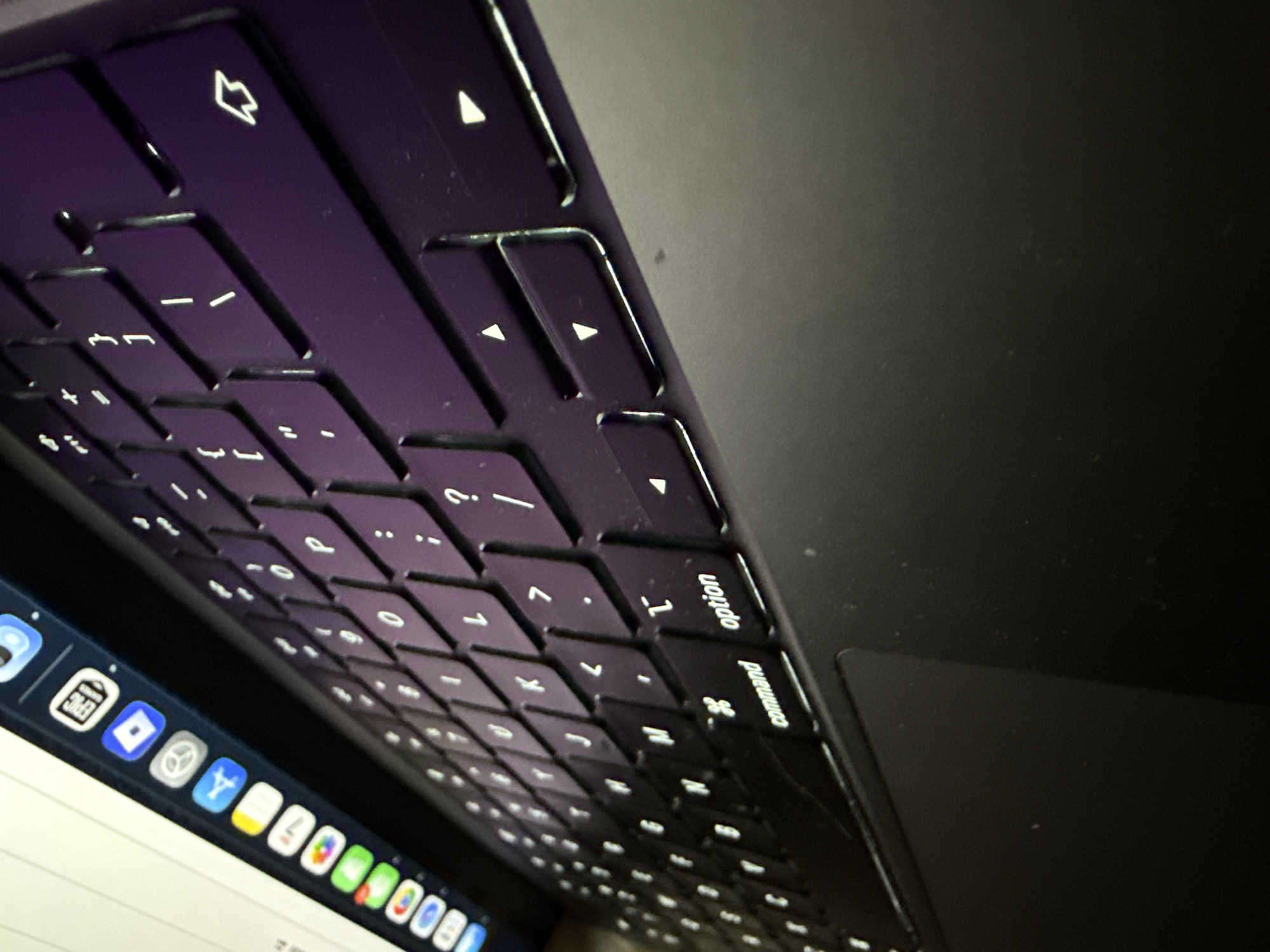 A close-up, low-angle photograph, taken indoors in the city of Kardzhali, Bulgaria, details a dark-colored laptop. The image prominently features the laptop's backlit keyboard and a portion of its screen. The keyboard's dark keys display illuminated white characters, which exhibit a distinct purple color cast attributed to ambient lighting conditions. Clearly visible keys include 'command,' 'option,' 'T,' '-', '/', '.', ',', and various directional arrow keys, along with the 'return' key. A dark, minimally reflective trackpad is situated to the left of the keyboard. The lower part of the laptop's screen is visible in the upper right, showcasing a bright white background with a row of colorful application icons in the dock, such as a web browser, mail, calendar, photos, Safari, App Store, settings, Roblox, and Epic Games. No individuals are present in the scene, and no specific environmental details beyond the laptop are discernible.