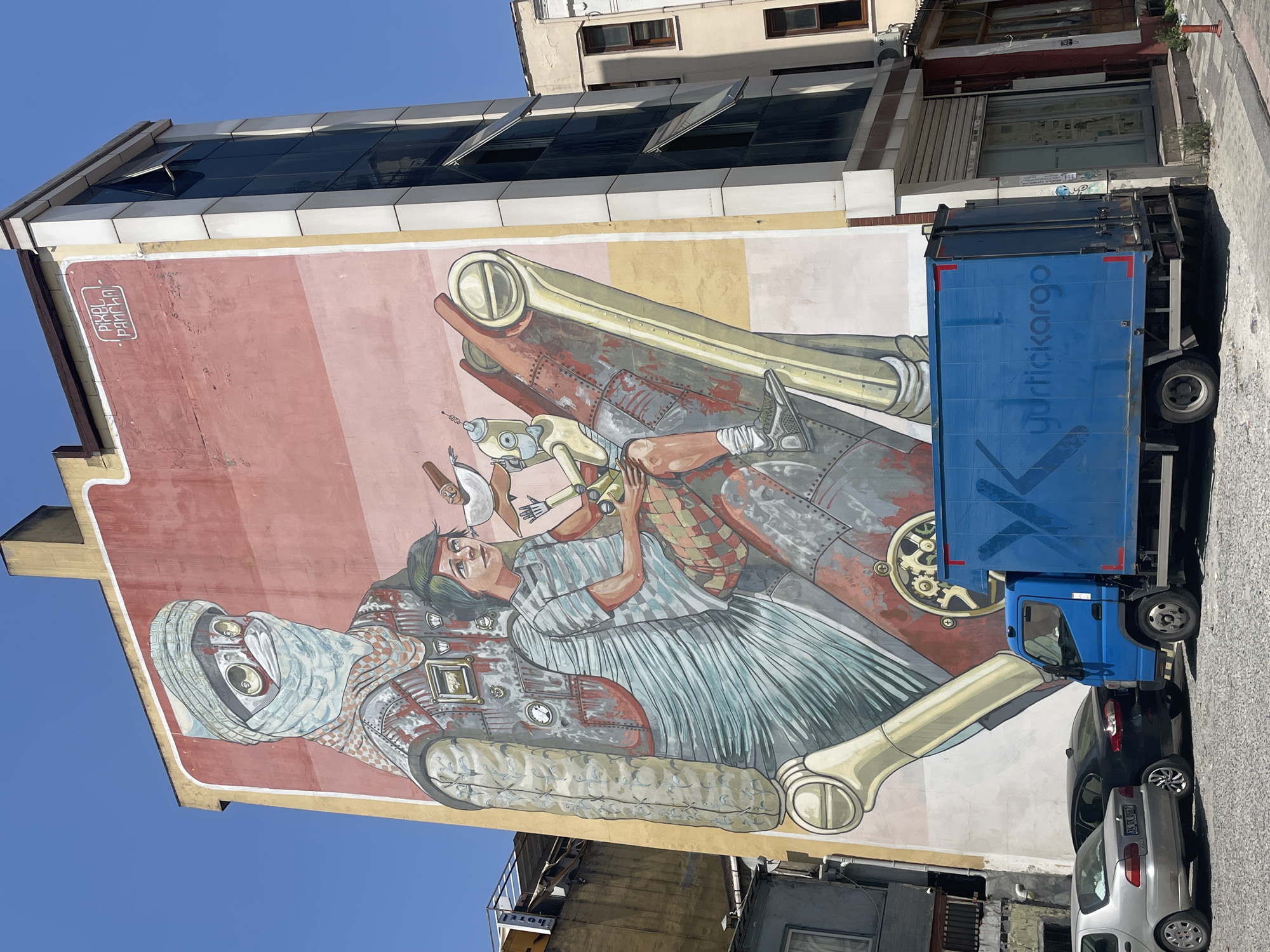 An outdoor mural adorns the side of a tall, multi-story building in Kadıköy, Turkey. The mural depicts a stylized, robotic figure wearing a headscarf and goggles, with a young person sitting on its shoulder. The robotic figure is detailed with metallic textures and gears, while the young person is depicted with a striped shirt and a checkered pattern on their shorts. The mural's background transitions from a pale yellow to a warm pink and red gradient. Above the mural, near the top right corner, the text "PIXEL PANCHO" is visible within a white border, indicating the artist.
In front of the building, a blue truck with the logo "yurtiçikargo" is parked on a gravel surface. To the left of the truck, two cars are parked: a silver sedan with a license plate partially visible and a black sedan. The environment appears to be a city street with other buildings and windows visible in the background. The sky is clear and blue.