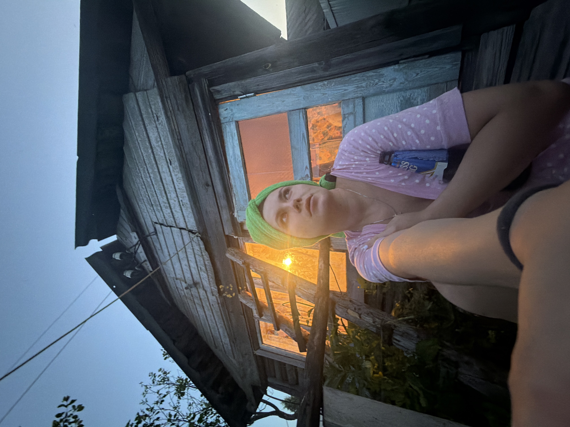 The image shows a young woman sitting on the steps of a rustic wooden structure, possibly a shed or an outhouse, in an outdoor setting. The structure appears weathered, with rough wooden planks and a damaged roof. A wooden ladder leans against the side of the structure, leading up towards the roofline. Through a window or opening in the structure, warm, orange light emanates, suggesting an interior light source. The woman is wearing a pink polka-dot robe and a green towel or wrap on her head. She is looking upwards and to the right, with a pensive expression. The background shows a clear sky with some branches of trees visible on the left. The lighting suggests it is either dawn or dusk, with the warm light from the structure contrasting with the cool blue of the sky. The location is identified as Красный Маяк, Russia.