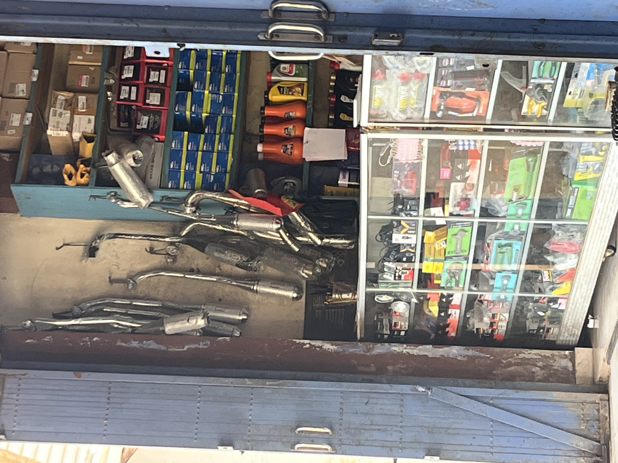 This is a medium shot, eye-level, exterior view of a motorcycle parts shop in Karang Sari, Indonesia. The shop features a blue metal door on the left and a blue metal frame for the shop interior on the right. The shop is filled with various motorcycle parts displayed on shelves and racks.
In the foreground, to the left, are several silver exhaust pipes hanging from hooks on the wall. To the right, a multi-tiered glass display cabinet showcases more motorcycle parts, including helmets, tools, and other accessories. Behind the glass cabinet, on shelves, are numerous boxes and containers of various sizes and colors, likely containing motorcycle parts and accessories. Several orange bottles and a yellow bottle are also visible on one of the shelves.
The overall impression is of a well-stocked motorcycle parts shop, ready to serve its customers. The lighting is natural, with a slightly overcast sky, and the scene appears to be during daytime. There are no people visible in the image.