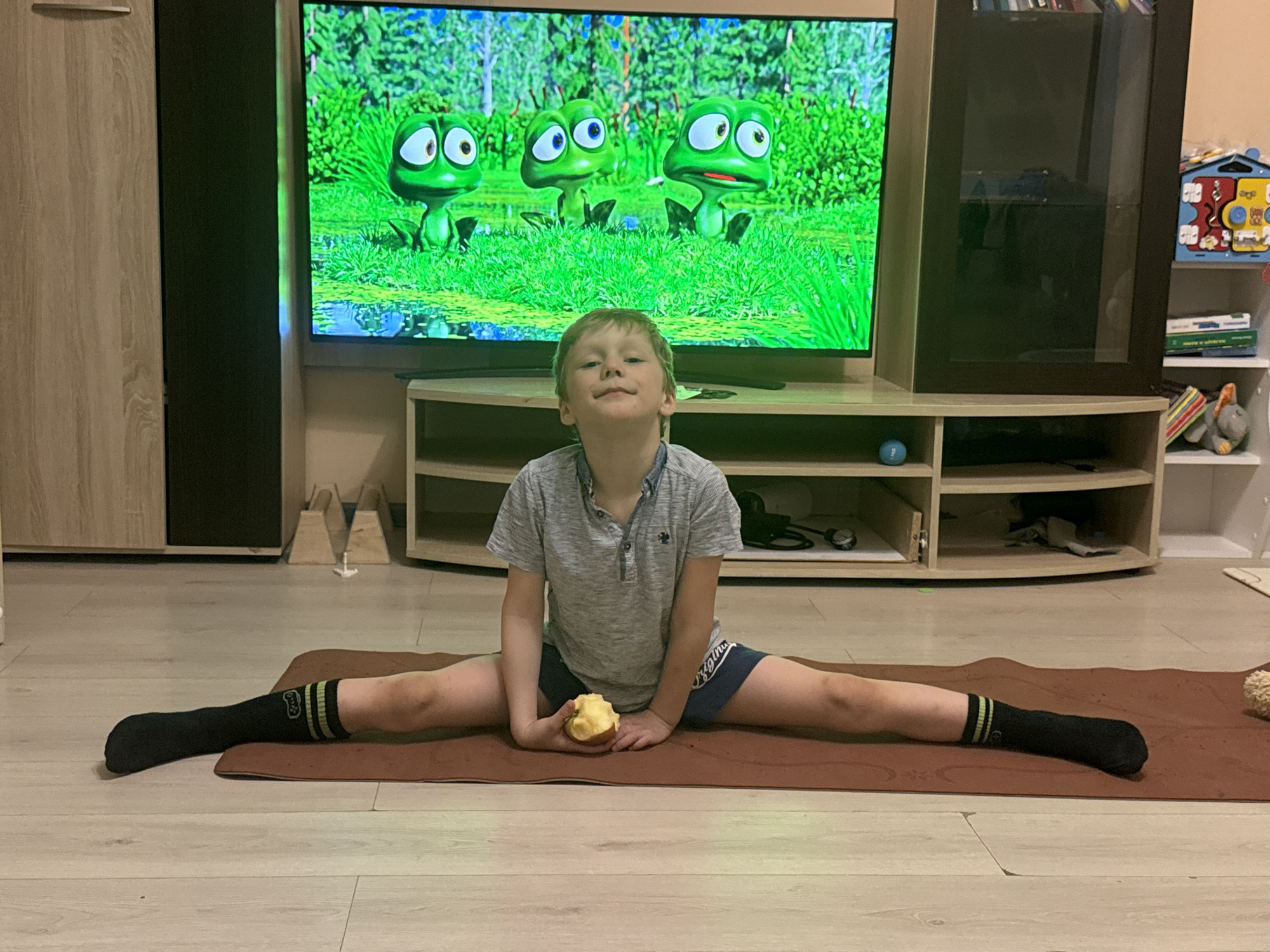 A young boy, appearing to be around 6-8 years old, is seated on a brown exercise mat in the middle of an indoor living space, likely a home in Tobolsk, Russia. He is performing a full split, demonstrating flexibility. He looks directly at the camera with a gentle smile, holding a partially eaten apple in his hands, suggesting he is taking a break or enjoying a snack during his exercise. He wears a grey short-sleeved collared shirt, blue shorts with the word "Originals" partially visible on them, and black socks with green and yellow stripes, which have a small, unidentifiable logo.
In the background, a large flat-screen television is displaying a vibrant animated cartoon featuring three green, anthropomorphic frog-like characters with large eyes, situated in a lush green natural environment with water and reeds. Below the television is a light-colored entertainment unit with open shelves, on which a blue ball and some cables are visible. To the left of the TV is a tall wooden cabinet, and to the right, a darker cabinet with a glass door. Further to the right, a white multi-shelf unit holds various items, including a colorful house-shaped toy and a grey stuffed elephant. The room has light-colored wooden or laminate flooring. The overall scene suggests a casual, active, and comfortable home environment.