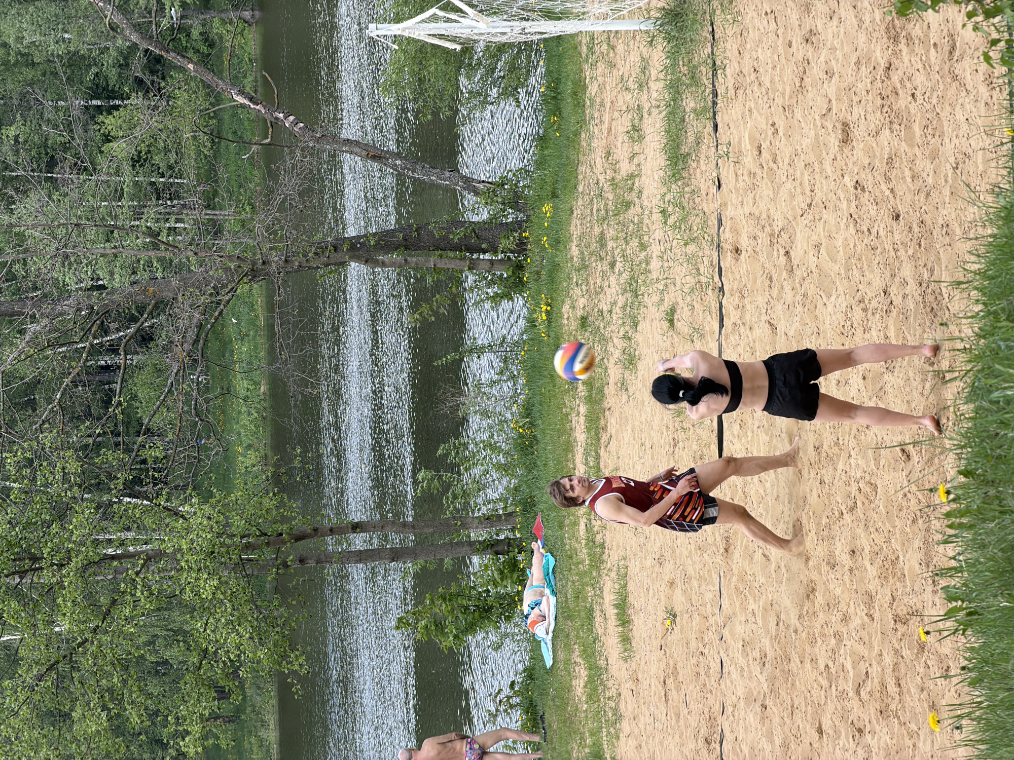 A horizontal medium shot depicts a scene at a beach volleyball court in Malakhovka, Russia, near a lake. Two individuals are actively engaged in a volleyball game. One person, dressed in a maroon and orange patterned tank top and dark shorts, is positioned lower on the court. The other person, wearing a black sports bra and black shorts, is positioned higher on the court, appearing to strike the volleyball. The volleyball is visible in mid-air. The playing area is sandy, delineated by a net supported by white posts. The lake runs along one side of the court, with a grassy area and trees separating the sand from the water. In the distance, on the grass near the lake, one person is sunbathing on a blue towel.