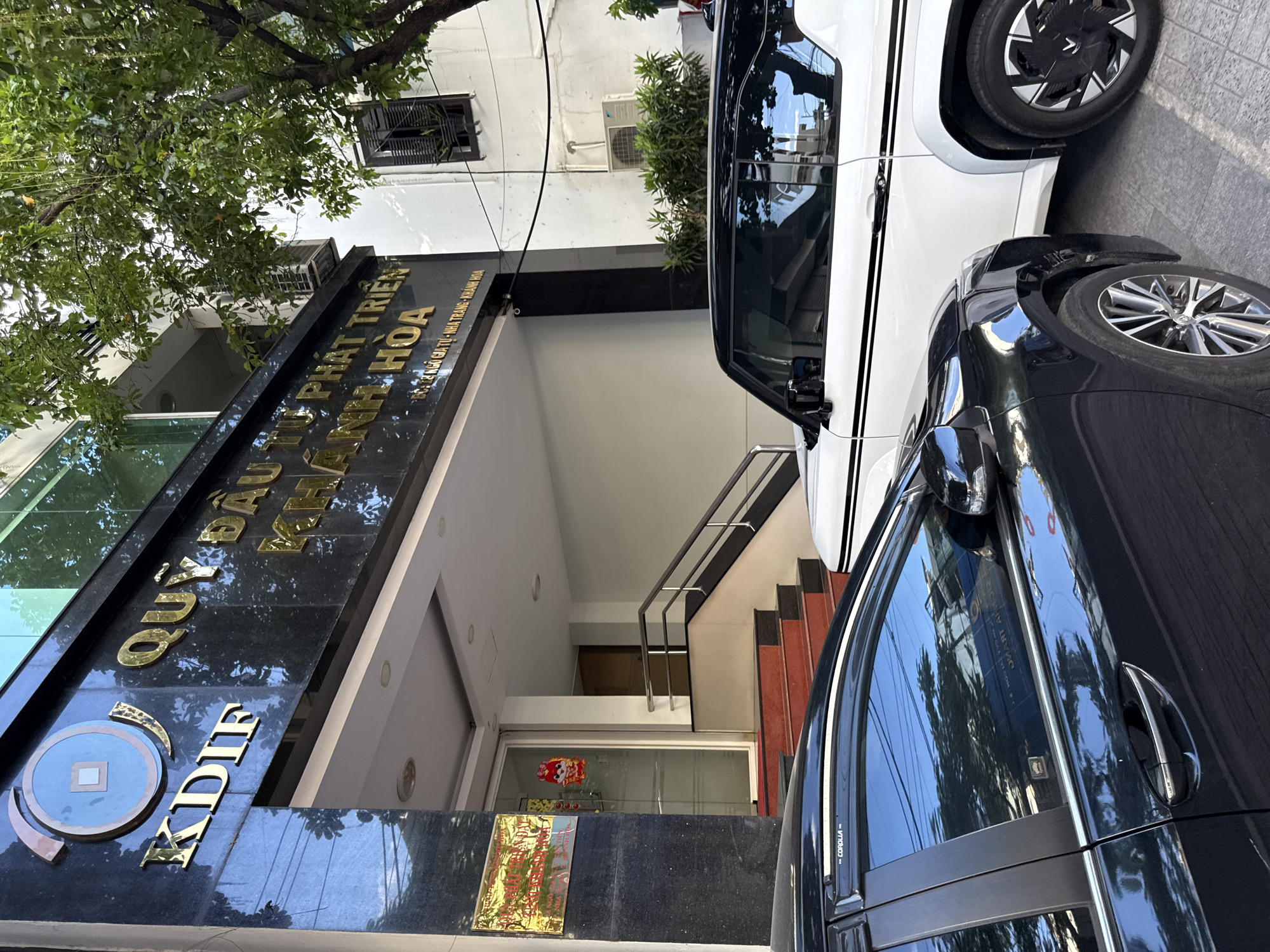 The image shows the entrance to a building with gold lettering that reads "K DIF QUỸ ĐẦU TƯ PHÁT TRIỂN KHÁNH HÒA" which translates to "K DIF Investment and Development Fund of Khanh Hoa". Below this, smaller text indicates the address: "Số 76 Ngô Gia Tự - Nha Trang - Khánh Hòa". This confirms the location is in Nha Trang, Vietnam.
The entrance is marked by a black marble facade with gold lettering. To the left of the entrance, a golden plaque is affixed to the wall. The entrance itself features glass doors with red and black steps leading up to it, and a metal railing alongside the stairs. Inside the entrance, a dimly lit hallway with a door can be seen.
Outside the building, two cars are parked in front. The car in the foreground is dark-colored, possibly black, with a visible "COROLLA" badge on the window frame. The car behind it is white with black detailing. A tree with green foliage is visible to the right of the building, partially obscuring the upper floors. An air conditioning unit is mounted on the exterior wall of the building. The overall scene depicts the exterior of a commercial building in an urban environment.
