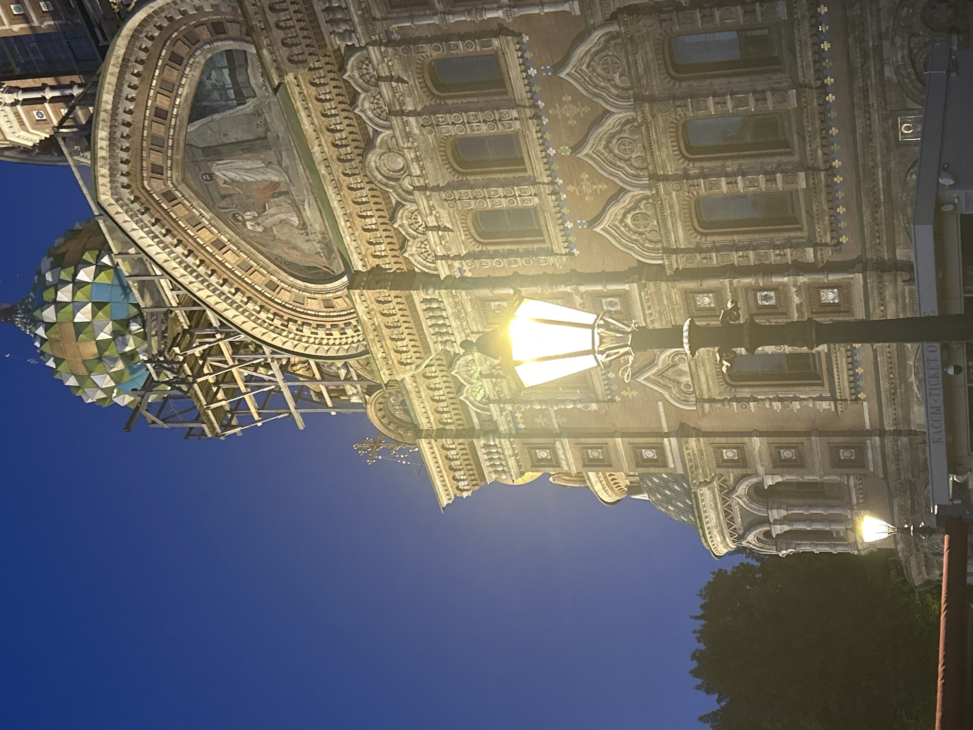 An external, low-angle, vertical shot captures the Church of the Savior on Spilled Blood in St. Petersburg, Russia, at dusk. The sky is a deep blue, transitioning from lighter hues at the horizon. The ornate, red-brick facade of the church, illuminated by streetlights, features intricate architectural details, including arched windows, mosaics, and decorative stonework. A prominent street lamp stands in the foreground, casting a warm glow on the scene. The church's iconic, multicolored onion dome is partially visible at the top, with scaffolding suggesting ongoing restoration work. To the left, a dense cluster of dark green trees provides a stark contrast to the illuminated architecture. Below the church's main facade, a sign with Russian text, likely indicating ticket sales, is visible. The overall atmosphere is one of historical grandeur and tranquility.
