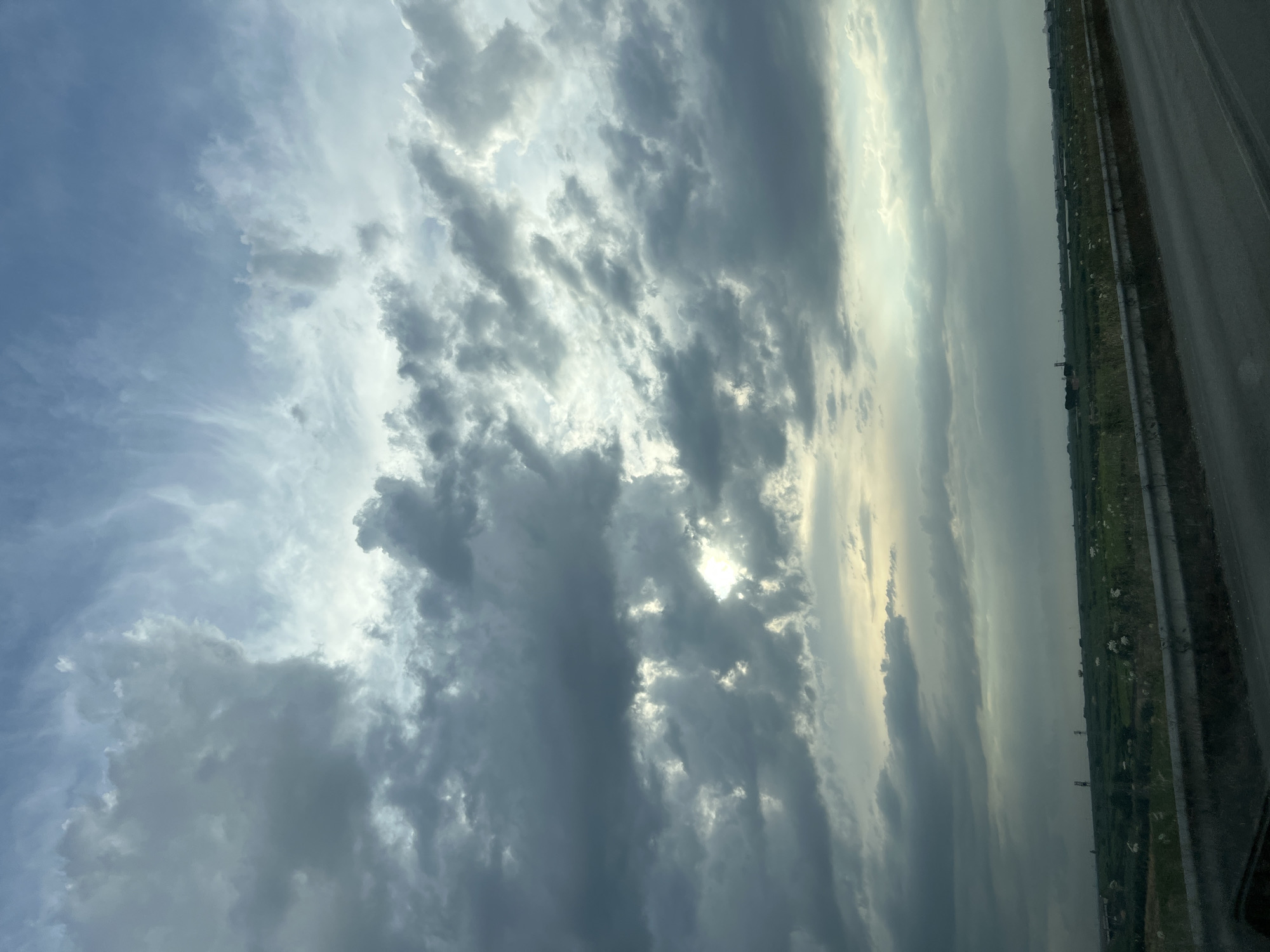 The image displays a panoramic view of the sky and landscape, likely taken from a moving vehicle on a road. The sky is filled with dramatic, overcast clouds, ranging in color from dark grey to lighter shades, with some patches of sunlight breaking through. The clouds appear dense and textured, suggesting impending or recent rainfall. Below the sky, a flat, green expanse of land stretches to the horizon. The landscape is dotted with what appear to be agricultural fields, some with visible white objects, possibly livestock or scattered vegetation. In the distance, several tall, slender structures, resembling utility poles or small towers, are visible. A concrete barrier runs along the edge of the road in the foreground, with a paved surface, presumably a highway or road, occupying the bottom portion of the image. The overall impression is of a rural or semi-rural setting under a moody, overcast sky. The location is identified as Barletta, Italy.