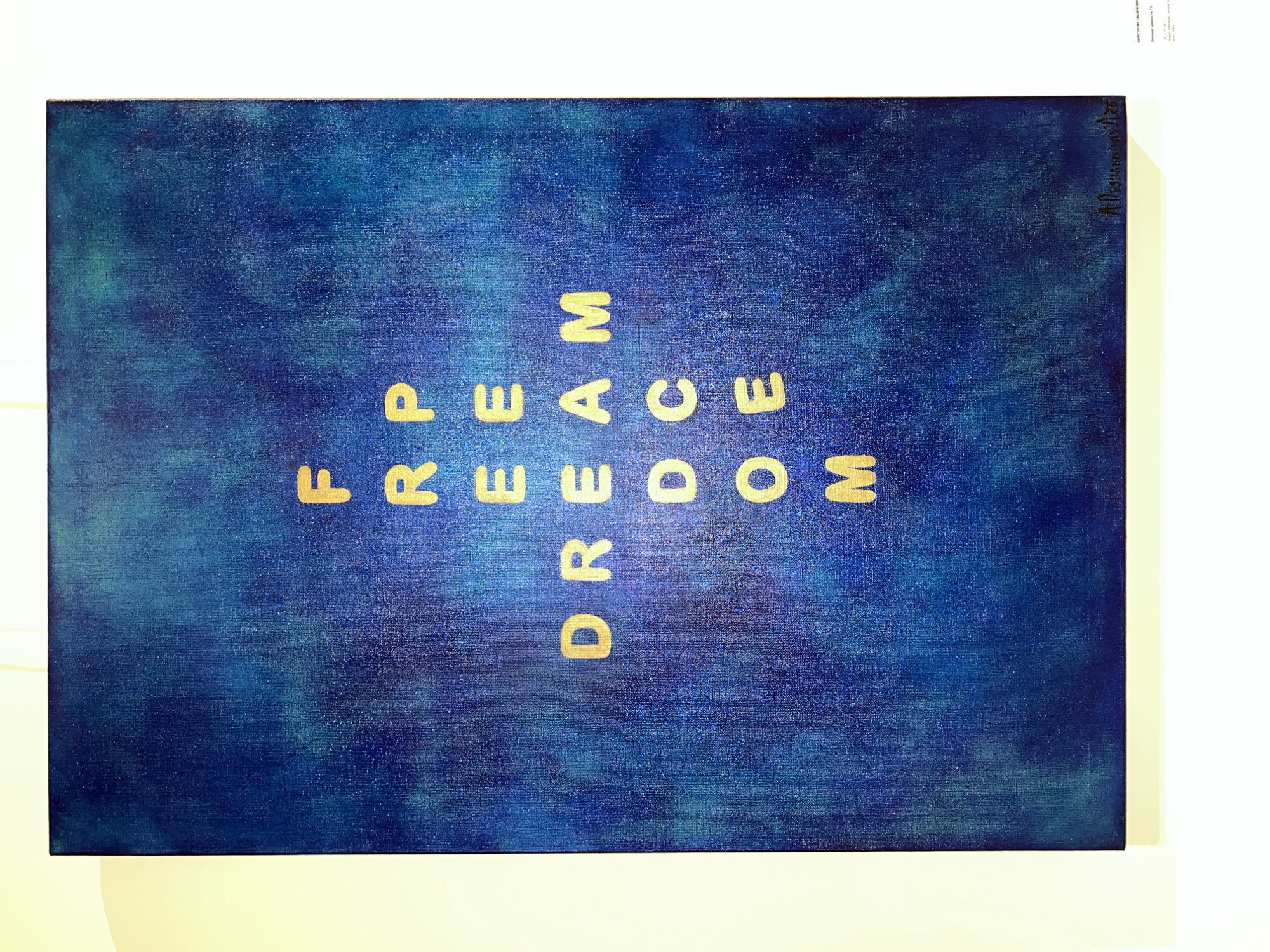 The image shows a dark blue canvas painting with the words "FREEDOM" spelled out vertically in gold lettering. The painting is on a white surface. There are markings in black ink in the upper right corner. The canvas is a rectangular shape. There is no additional information provided in the image about the location of the city of Moscow, Russia.