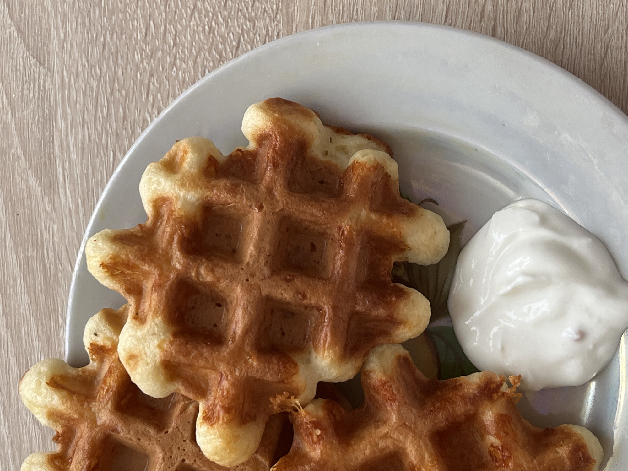 A high-angle, close-up shot shows a plate of waffles with a dollop of white cream on a wooden surface. The waffles are golden-brown and arranged irregularly on the plate. The white cream is smooth and glossy. The plate is predominantly white with hints of other colors. The wooden surface has a light brown color. There is no discernible information in the image that allows for determining its location in Chaikovsky, Russia, or any other specific location. No people are visible.