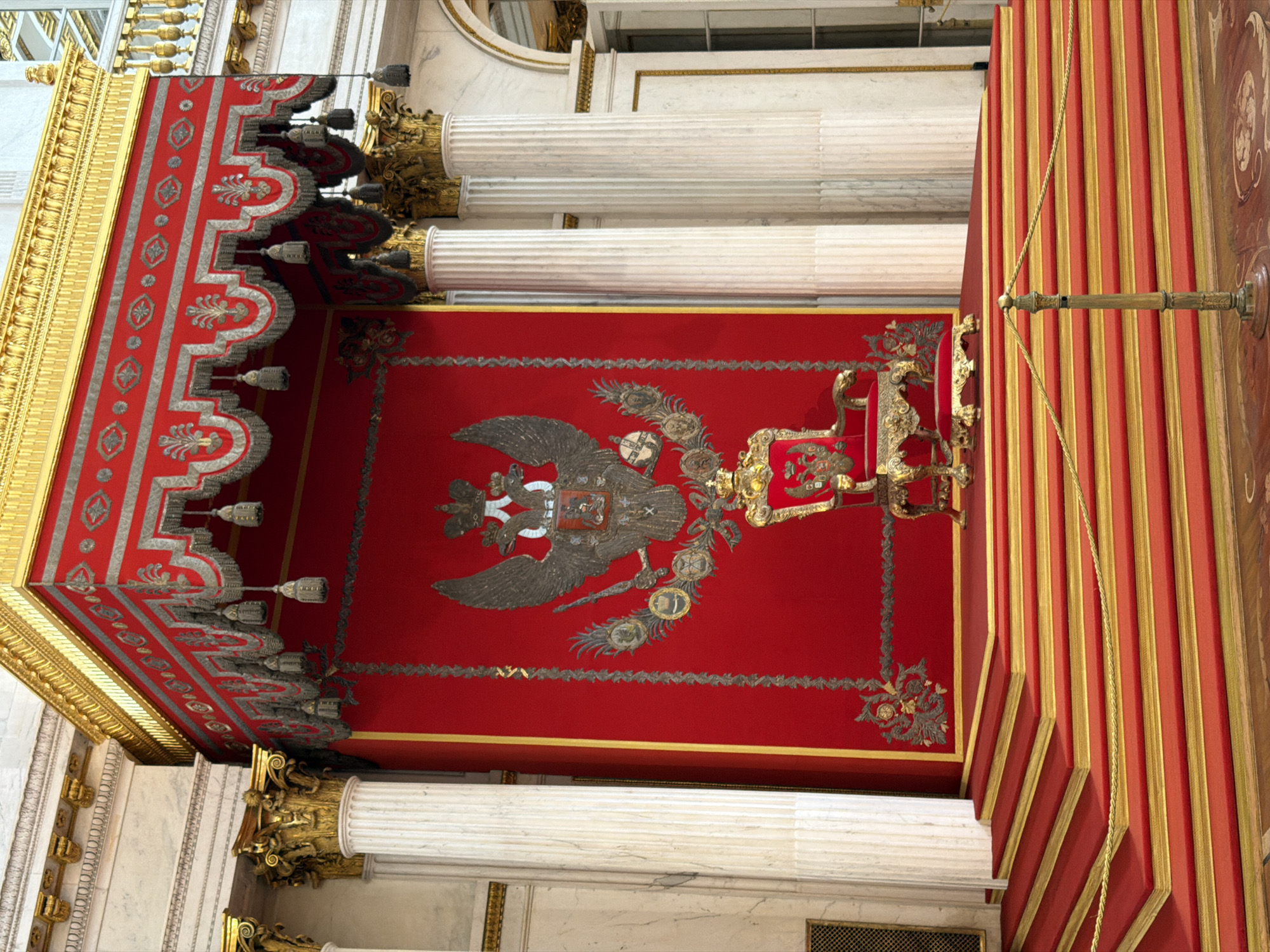 This is a photograph of the interior of a grand building, likely a palace or cathedral, in Saint Petersburg, Russia. The image is oriented vertically, showing a high ceiling with ornate red and gold decorations. A prominent feature is a large, dark double-headed eagle emblem, possibly a coat of arms, set against a vibrant red background on the ceiling. The eagle is detailed, with intricate feathering and what appears to be a shield on its chest.
Surrounding the central emblem are decorative borders and elements, including gold and silver trim. The ceiling is further embellished with elaborate golden architectural details, including what look like decorative tassels hanging from a gilded fringe. Large white fluted columns, topped with ornate gold capitals, rise towards the ceiling, framing the view.
The overall impression is one of opulence and grandeur, typical of Russian imperial architecture. The lighting appears to be artificial, suggesting the photograph was taken indoors during the day or evening. The context of Saint Petersburg, Russia, points to a historically significant and architecturally rich location. There is no visible text in the image. The scene conveys a sense of solemnity and historical significance, with no people or specific activities visible, focusing entirely on the architectural and decorative elements.
