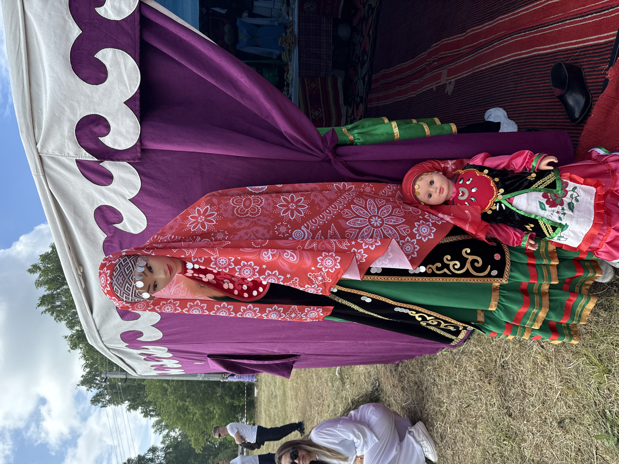 In the city of Nurkeevo, Russia, a mannequin and a doll are displayed in front of a tent. The mannequin is dressed in a traditional red headscarf with silver accents and a colorful, layered dress. The doll, also dressed in traditional attire, is positioned at the mannequin's feet. The tent has a purple fabric interior with a decorative trim. The scene is set outdoors on a grassy area, with trees and a blue sky visible in the background. Several people are present in the background, some of whom appear to be visitors. A stall with goods is visible inside the tent.