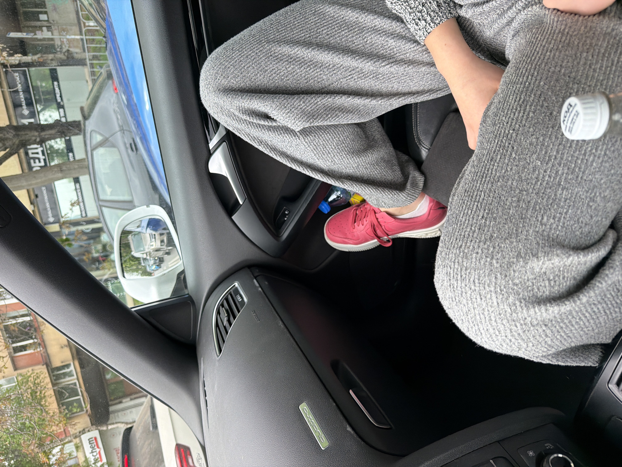 The image shows the interior of a car, specifically the front passenger side. A person is seated, wearing gray sweatpants and a gray knitted top, and pink sneakers. A bottle of water is visible near the person's leg. The dashboard and interior elements of the car are visible, including an air vent and a "quattro" emblem. Through the window, buildings and signs written in Cyrillic can be seen. Based on the Cyrillic script and the presence of a "Balkan Finance" sign, the location is identified as Sofia, Bulgaria.