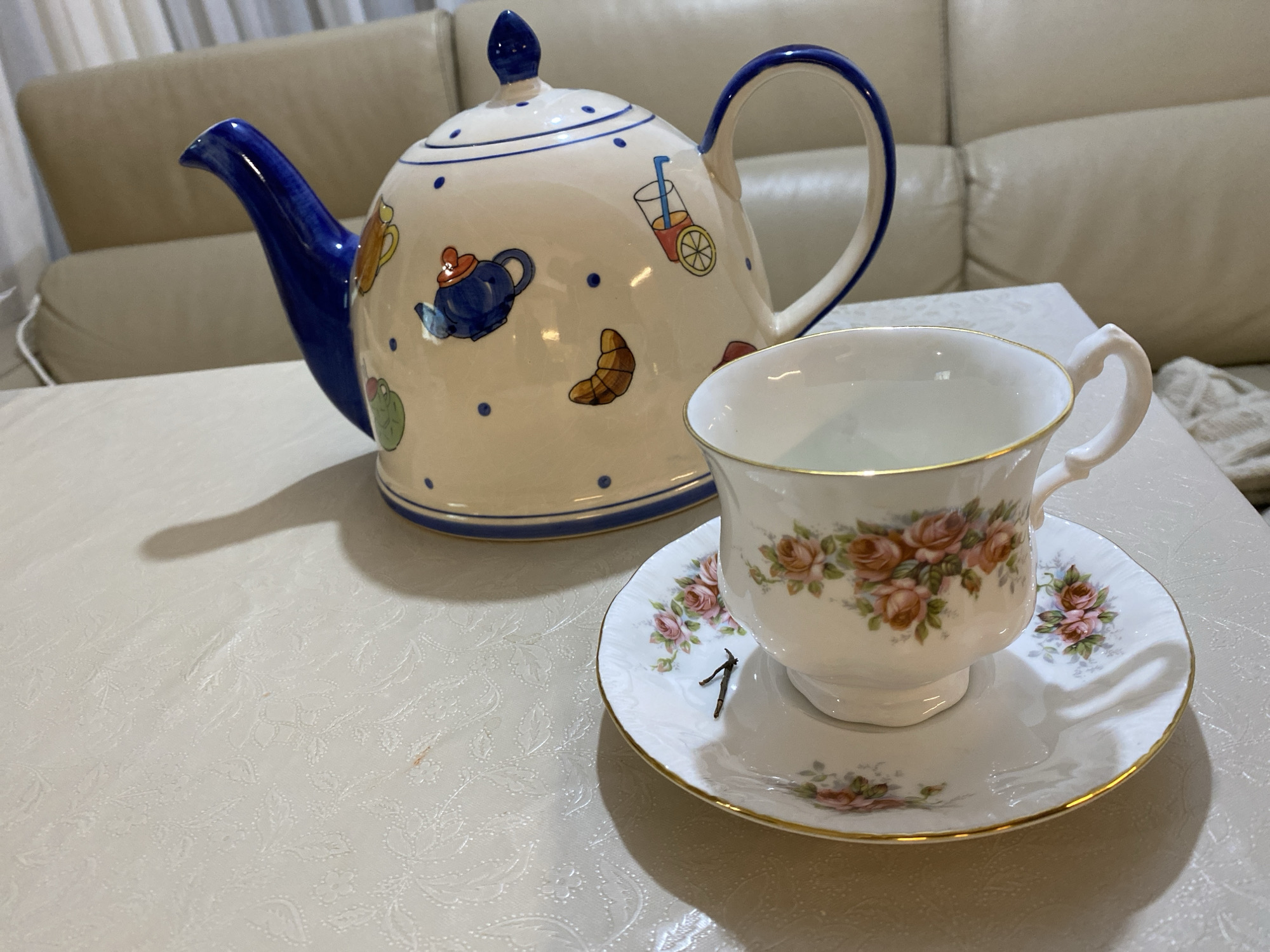 A still life on a light-colored, patterned tablecloth features a teapot and a teacup with a saucer. The teapot is cream-colored with blue trim and decorative illustrations of various items such as a teapot, croissant, and glass of juice. The teacup and saucer are white with a floral rose pattern and gold trim. There is a dark thin object on the saucer, resembling the number "1". The setting appears to be indoors, with a light-colored couch visible in the background. There is no indication of Budva, Montenegro.