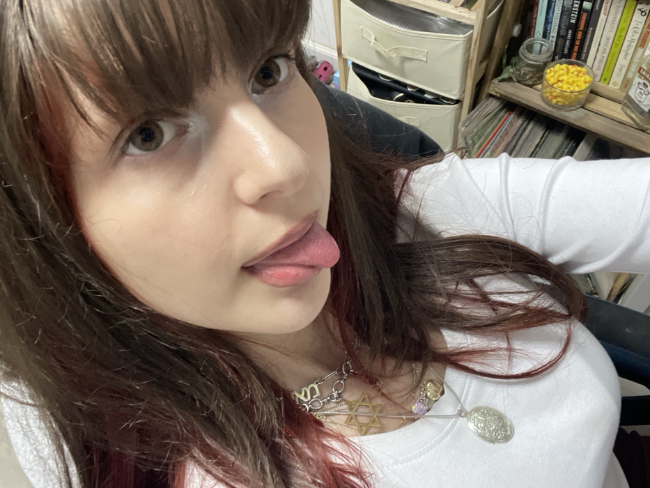 A woman with brown hair and bangs, wearing a white long-sleeved shirt, poses close to the camera. She has light skin, brown eyes, and her tongue is sticking out. She is wearing multiple necklaces with various pendants, including a Star of David and a locket. Behind her, there's a shelf with books, storage boxes, and a jar filled with yellow candies.