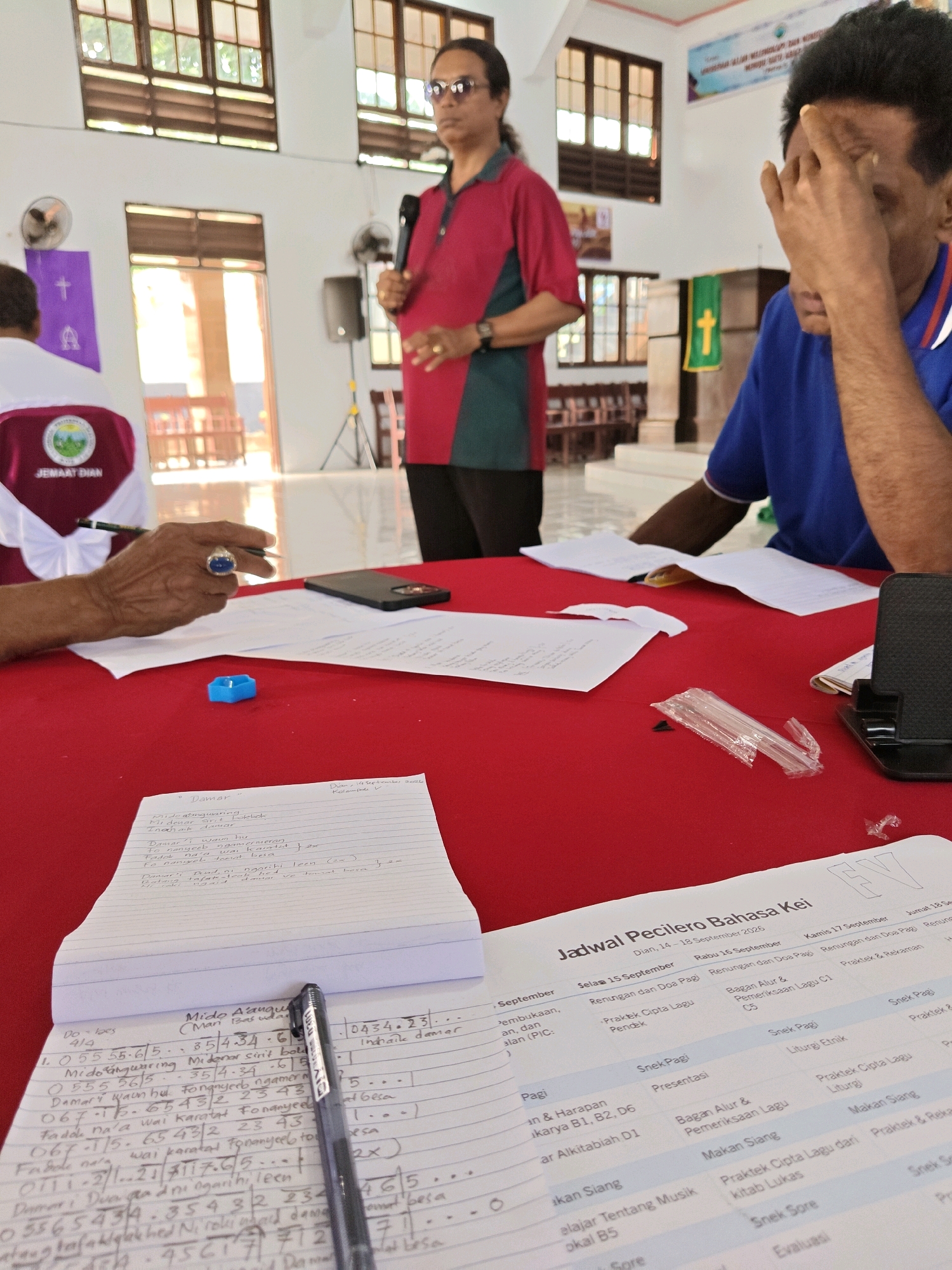 The image depicts an indoor gathering, likely a church service or meeting, at the Dian Darat community hall. In the foreground, a red table is laden with papers and a notebook. A hand, adorned with a large blue ring, holds a black pen, seemingly in the process of writing. A black smartphone rests on the table, alongside scattered paper scraps. Further back, a person in a blue shirt is seated, with their hand covering their forehead. To the left, another person in a maroon and green shirt stands, holding a microphone and speaking. Behind them, a purple banner with a white cross is visible. The room features wooden window frames and the overall ambiance suggests a casual, community-focused event. The note at the top right of the notebook indicates "Dian, 14 September 2026, Kelompok V," placing the setting within the Dian Darat area on this specific date and context.