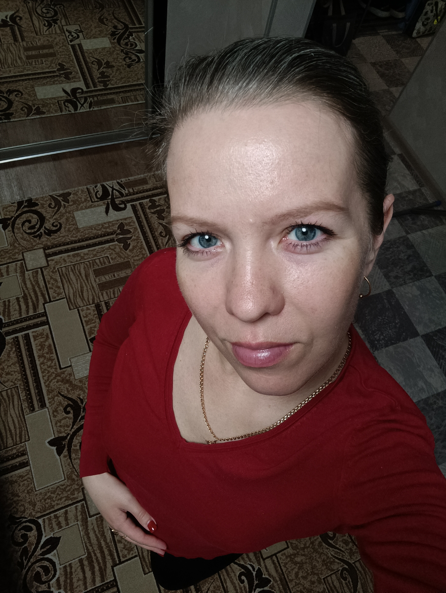 A high-angle, close-up indoor selfie features a woman looking directly towards the camera. She has light skin, bright blue eyes, and dark hair pulled back, revealing some grey strands. Her lips are a glossy light pink. She wears a red long-sleeved top with a scoop neckline and a gold-colored chain necklace. A small gold hoop earring is visible on her right ear, and a ring is on her left ring finger, with her nails painted red. Her left hand rests gently on her lower torso.
The background shows a patterned floor covering, likely linoleum or carpet, featuring intricate geometric and curvilinear designs in shades of beige, brown, and dark brown. This pattern transitions to a dark grey and black checkered design towards the upper right. On the left side, a dark, reflective vertical surface, possibly a wardrobe or mirror, shows a reflection of the patterned floor and a horizontal metallic strip. The lighting appears to be artificial. The setting is an indoor space, consistent with a residence in Chaikovsky, Russia. There is no visible text in the image.