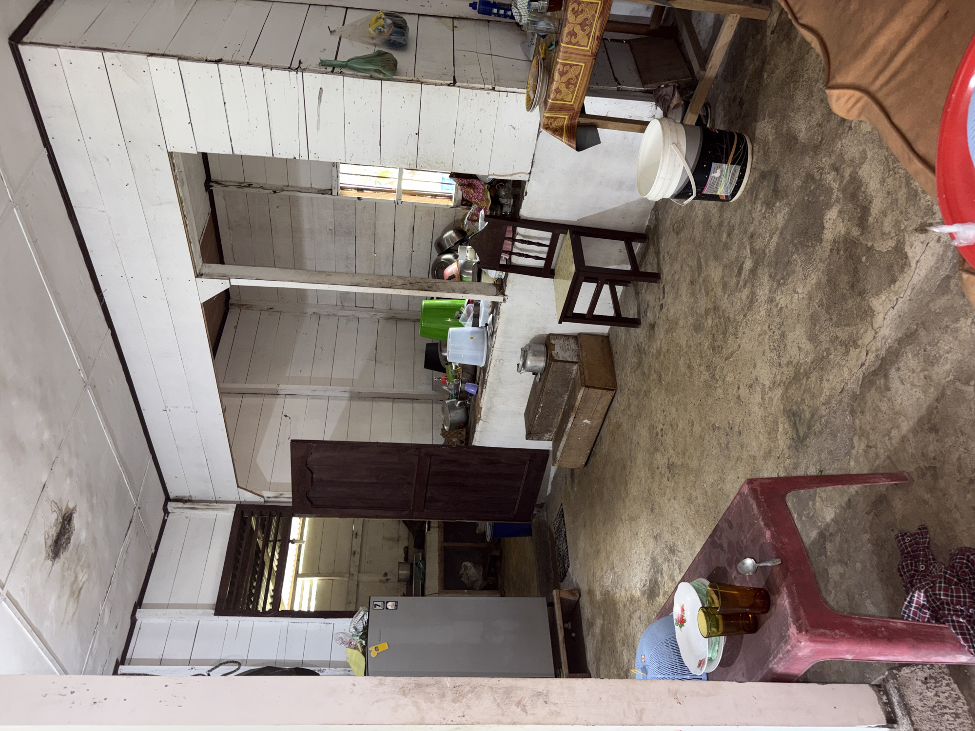 The image shows the interior of a rustic kitchen in Beunyot, Indonesia. The room has white, planked walls and a white, textured ceiling with some water stains. The floor is made of concrete and appears worn.
In the foreground, a red plastic table holds two amber glasses, a plate, and a spoon. To the left, a silver refrigerator stands on wooden blocks. A dark wooden door is open to reveal a glimpse of a kitchen area with countertops and cabinets.
In the middle ground, a dark wooden chair sits near a counter. There are various kitchen items on the counter, including pots, bowls, and containers. A wooden table with a patterned tablecloth is situated to the right, with plates and glasses on it.
In the background, a window with slats allows light to enter the room. Bags hang from a hook on the wall. A large white bucket with black markings is on the floor. The overall impression is of a lived-in, functional kitchen.