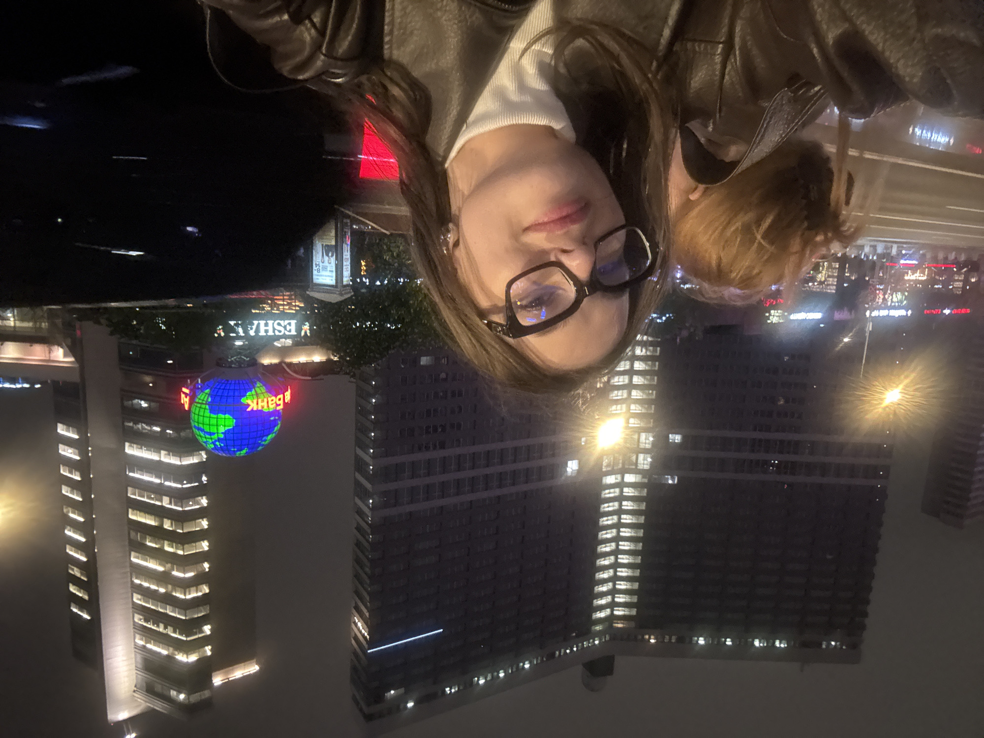 A selfie taken at night shows a woman wearing a brown leather jacket and glasses, with a light-colored top underneath the jacket. Behind her are city lights and buildings, including one with a large globe on top displaying continents. In the background, the words "ESHA", and "БАНК" can be partially seen. The location appears to be in Moscow, Russia due to the globe atop the building being the headquarters of MDM Bank.