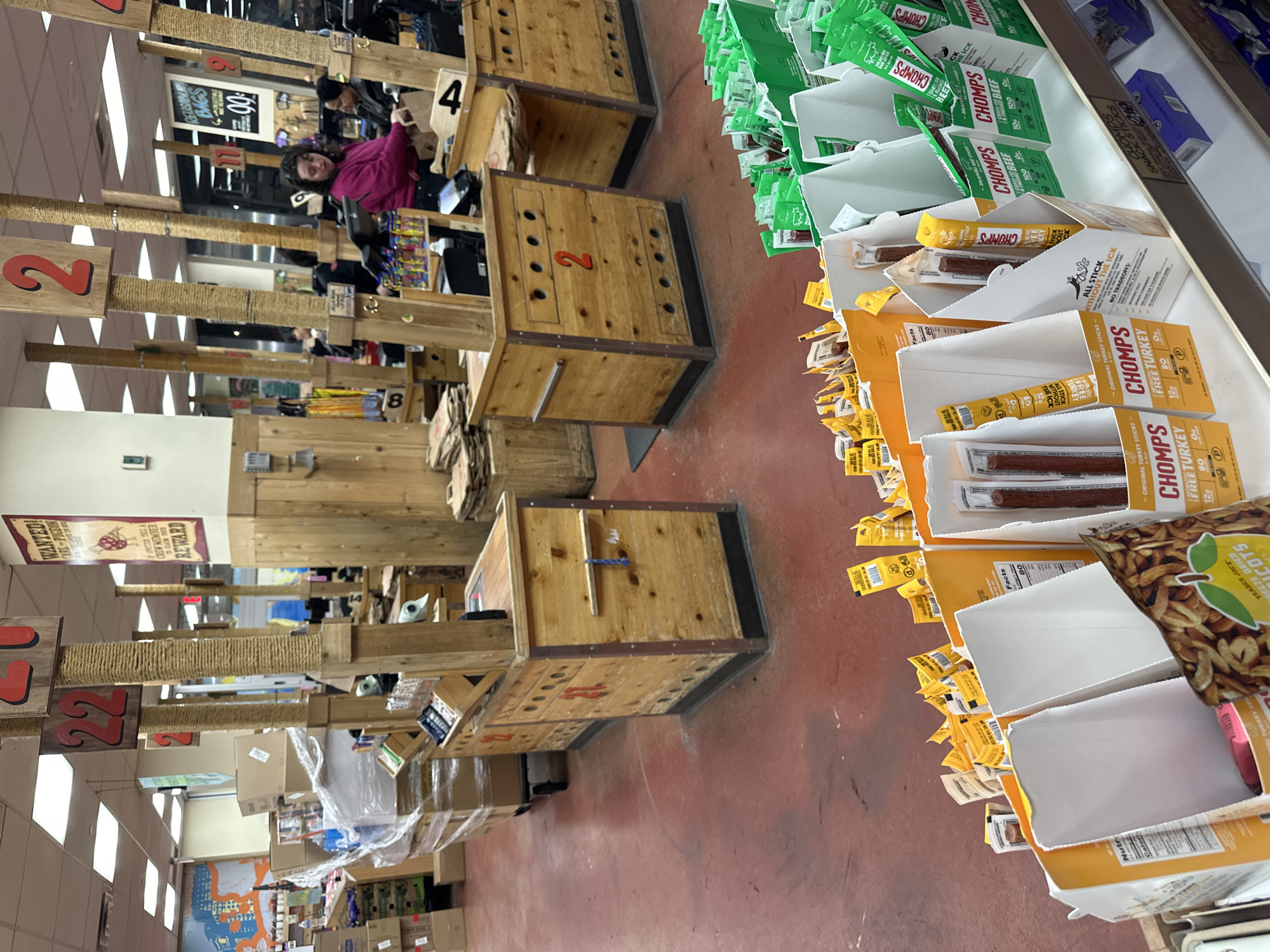 This is an indoor shot of a Trader Joe's store in New York City, United States. The camera is focused on the checkout area, which is set up with wooden booths numbered to guide customers. A person with dark hair wearing a pink jacket is at one of the booths. The shelves next to the checkout lanes are stocked with snack items, including boxes of "CHOMPS" brand meat sticks and bags of dried apricots. The floor is red. The overall atmosphere is that of a busy grocery store.