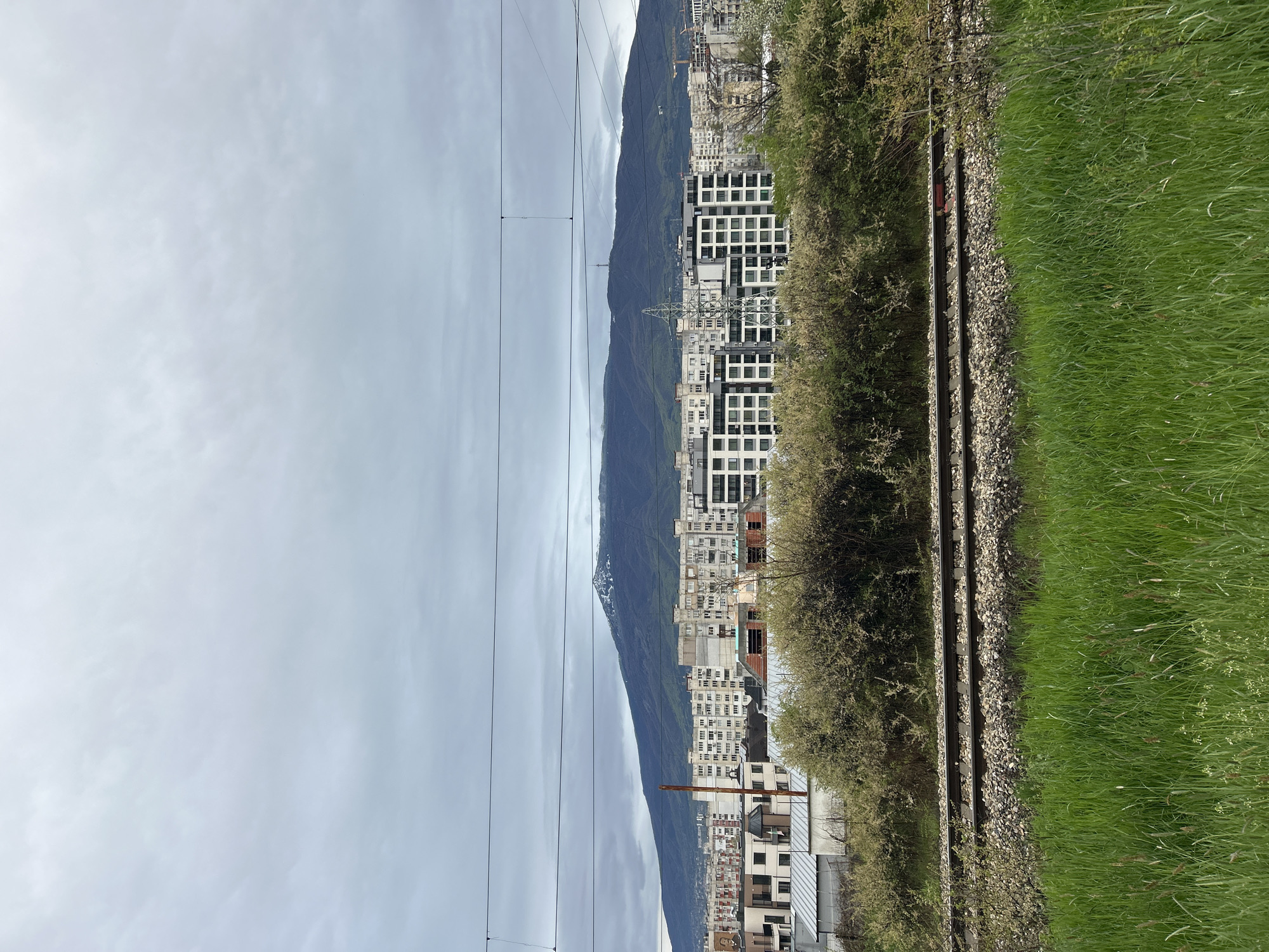 The image presents a wide, panoramic view of an urban landscape in Sofia, Bulgaria, set against a backdrop of a mountain range on an overcast day.
In the foreground, a pair of train tracks runs horizontally across the bottom section of the frame, laid on a bed of grey gravel ballast. To the immediate right of the tracks, a lush, vibrant green grassy embankment stretches out, while to the left, a line of dense, green bushes and trees partially screens the city beyond.
The midground is dominated by a sprawling city composed of numerous multi-story buildings. These structures display a mix of architectural styles, including modern blocks with repetitive window patterns and older or partially constructed buildings. An electricity pylon is visible within the urban sprawl, and multiple overhead power lines traverse the sky above the city. A slender telecommunications tower can be faintly seen on a lower part of the distant mountain.
The background features a large mountain range, appearing dark and rugged under the heavy cloud cover. Some higher elevations of the mountains show patches of white, possibly remnants of snow or exposed rock. The sky above is uniformly grey and cloudy, indicating an overcast day without direct sunlight.
No people or specific activities are depicted in the static scene. The image captures the confluence of urban development, infrastructure (train tracks, power lines), and natural mountainous terrain. There is no visible text in the image.