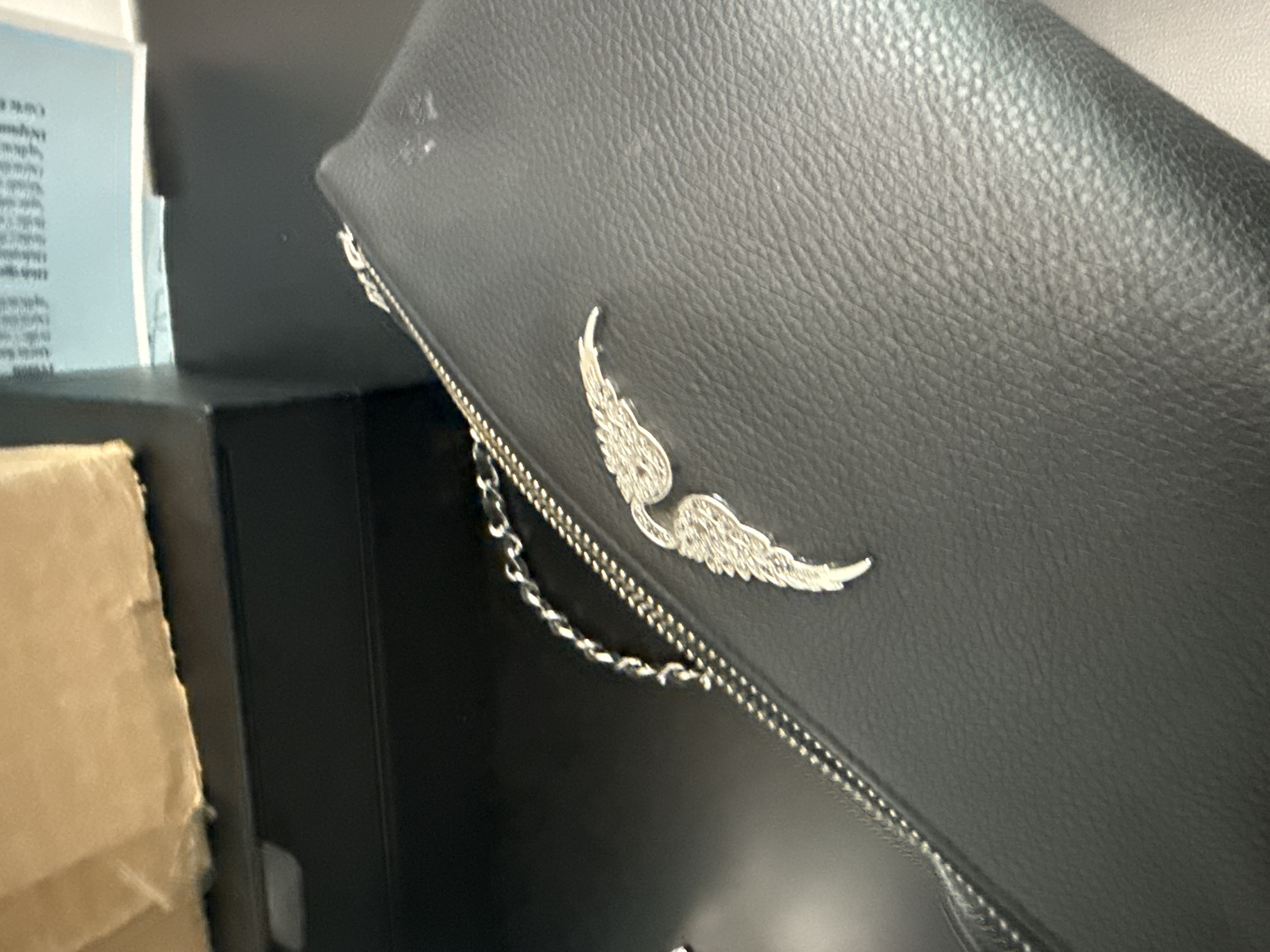 The image shows a close-up of a black leather bag. The bag has a silver wing emblem and a silver chain. The bag is next to a black box. The image was not taken in San Bernardino Tlaxcalancingo, Mexico.