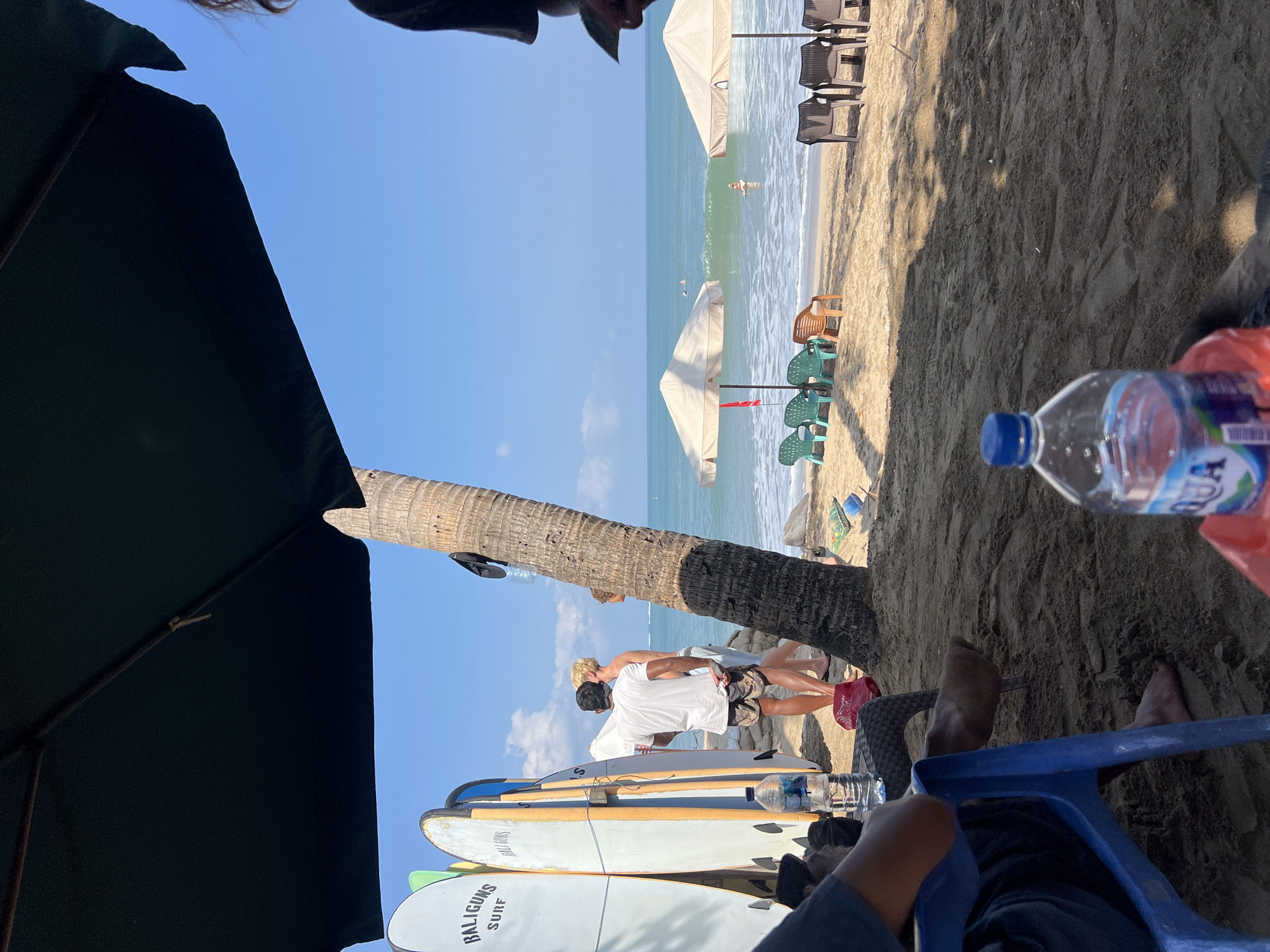 The image depicts a beach scene in Kuta, Indonesia. In the foreground, a person sits in a blue plastic chair on the sand, with their bare feet visible. To the left, a row of surfboards stands upright, some with "BALIGUNS SURF" branding. A palm tree trunk is prominent in the midground. Several beach chairs and umbrellas are set up on the sand, facing the ocean. The ocean is calm with small waves, and a few people are visible in the water, some appearing to be surfing. The sky is clear and blue.