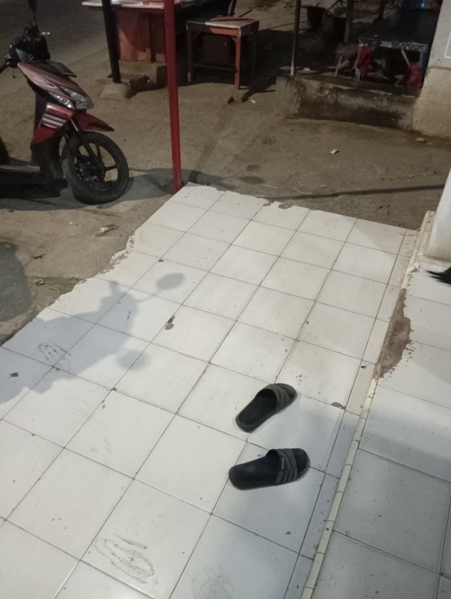 A red scooter is parked on a concrete surface next to a tiled walkway. Two dark-colored slides are placed on the tiled walkway. A thin, vertical red pole is present to the right of the scooter. In the background, there are structures that appear to be part of an outdoor market or roadside establishment. The scene is dimly lit, suggesting it is nighttime. The location is identified as Sliyeg, Indonesia.