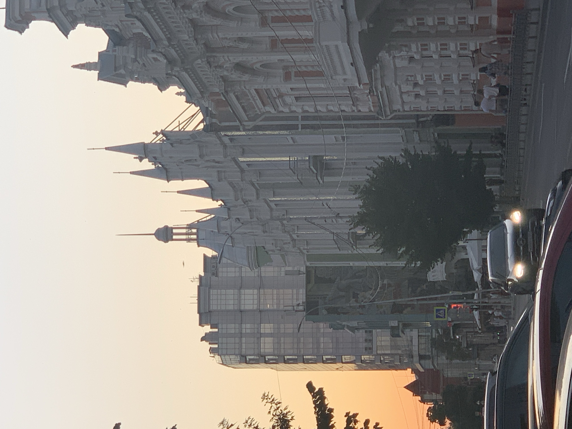 The image depicts a street scene in an urban environment during what appears to be sunset, with an orange glow in the sky. On the right side of the frame, an ornate, multi-story building with white and red architectural details and numerous windows is visible. To the left of this building is another older building with a more uniform facade and several spires, some of which are topped with pointed finials. Further to the left, a modern, tall glass-fronted building stands out. A tree with dark foliage is positioned in the middle ground, obscuring part of the older buildings. Several cars are on the road in the foreground, with one car's headlights illuminated. Pedestrians can be seen walking on a sidewalk behind a metal railing. A street sign with a pedestrian symbol and traffic lights are also present. The overall impression is a street with a mix of architectural styles, bathed in the warm light of dusk.