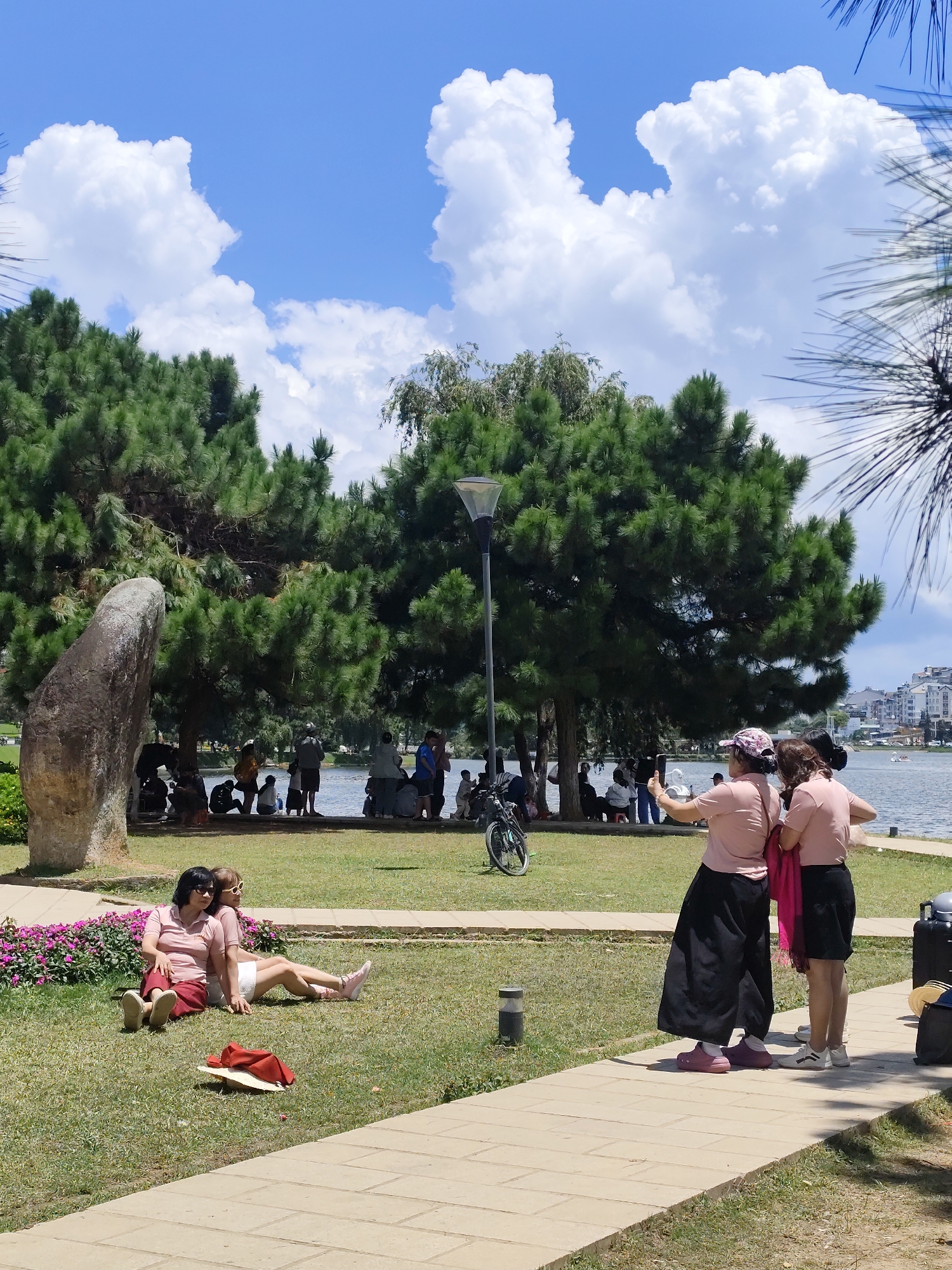 In Da Lat, Vietnam, this image captures a sunny day at Xuan Huong Lake. In the foreground, two women in pink tops and dark skirts stand on a paved path, one taking a selfie with her phone. To the left, on a grassy area beside blooming purple flowers and a large rock, two other women in pink tops and skirts relax, one lying down with her legs extended, the other sitting up. Further back, a group of people are gathered near the edge of the lake, with pine trees and a lamppost visible. In the background, buildings line the opposite shore of the lake, and a bicycle is parked on the grass. The sky is bright blue with scattered white clouds.